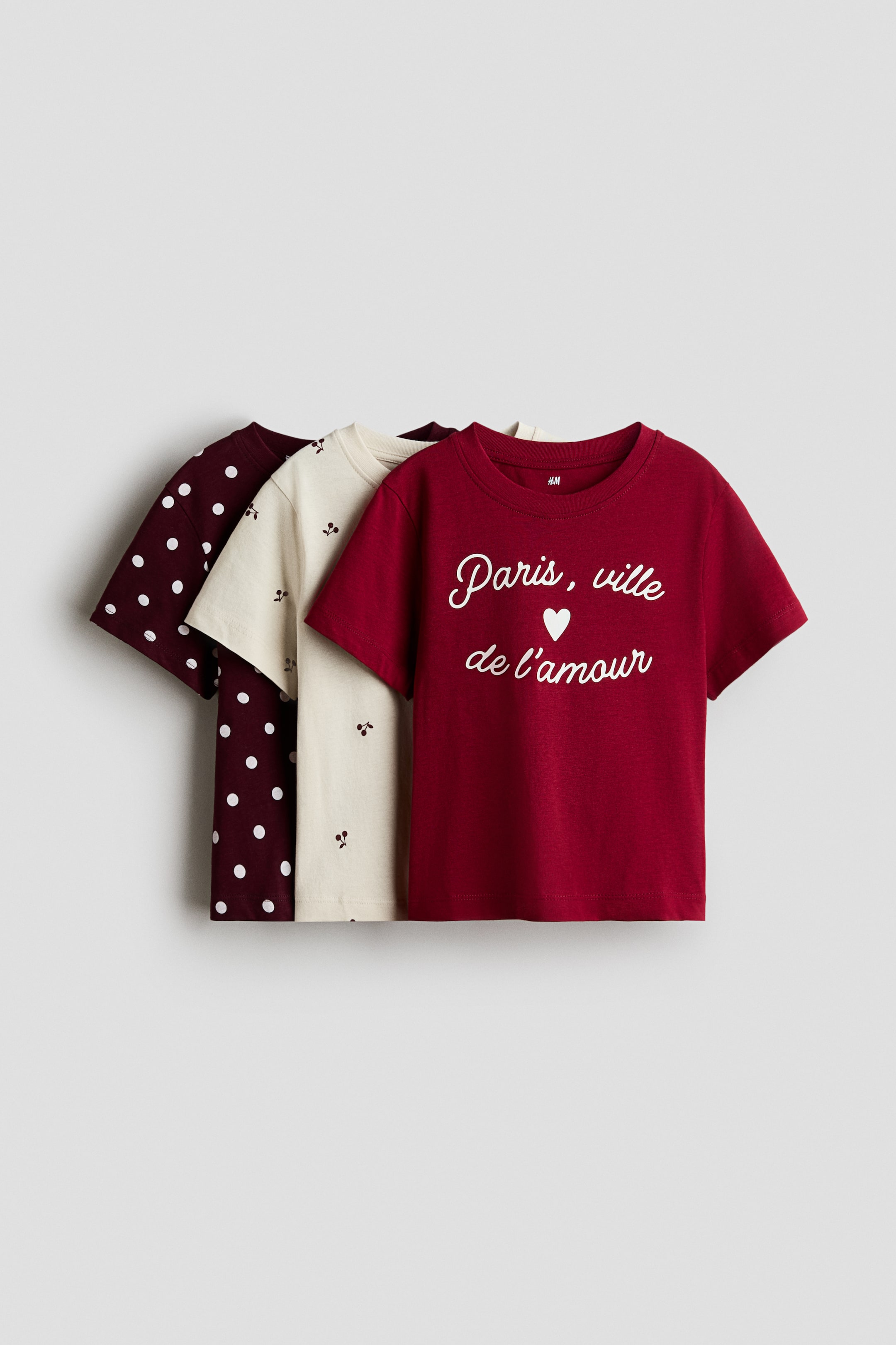 View larger image: Three short-sleeved cotton jersey t-shirts with round, rib-trimmed necklines are shown facing front in a layered presentation. The front t-shirt is dark red, featuring a white cursive graphic that reads "Paris, ville de l'amour" with a small heart, layered over a cream t-shirt with a scattered cherry pattern and a dark burgundy t-shirt with white polka dots.
