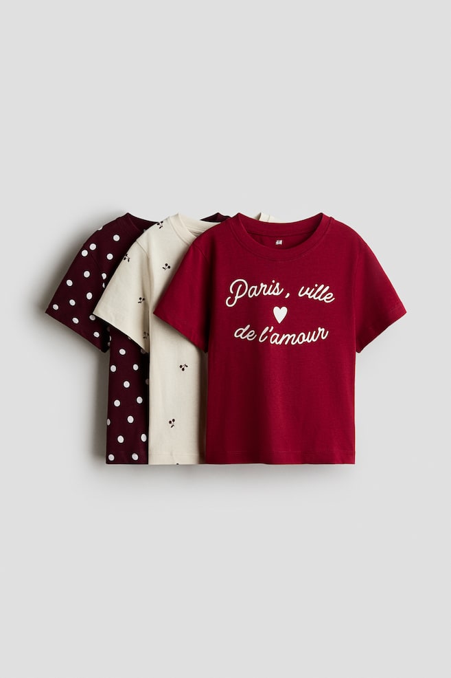 Three short-sleeved cotton jersey t-shirts with round, rib-trimmed necklines are shown facing front in a layered presentation. The front t-shirt is dark red, featuring a white cursive graphic that reads "Paris, ville de l'amour" with a small heart, layered over a cream t-shirt with a scattered cherry pattern and a dark burgundy t-shirt with white polka dots. - 1