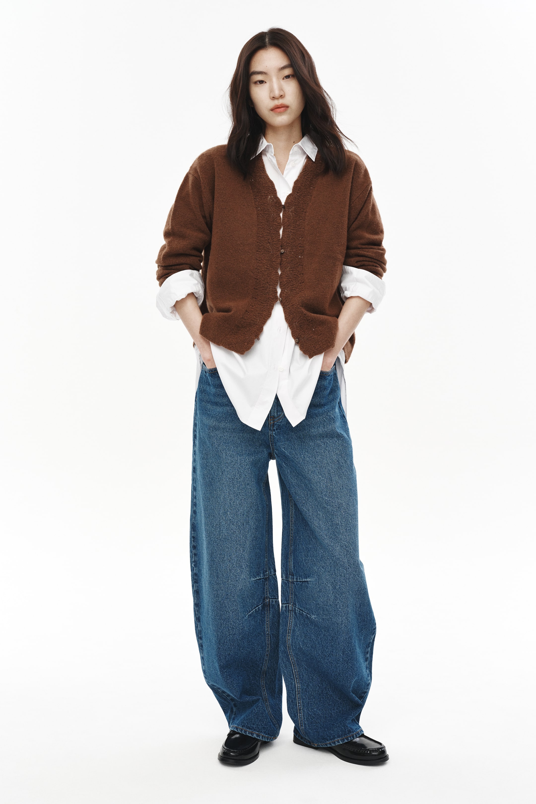 拡大画像を表示: 茶色のふわふわしたVネックカーディガンが白い襟付きシャツの上に着られ、膝の縫い目が特徴的なワイドレッグの青いジーンズが黒いローファーと共に着用されています。