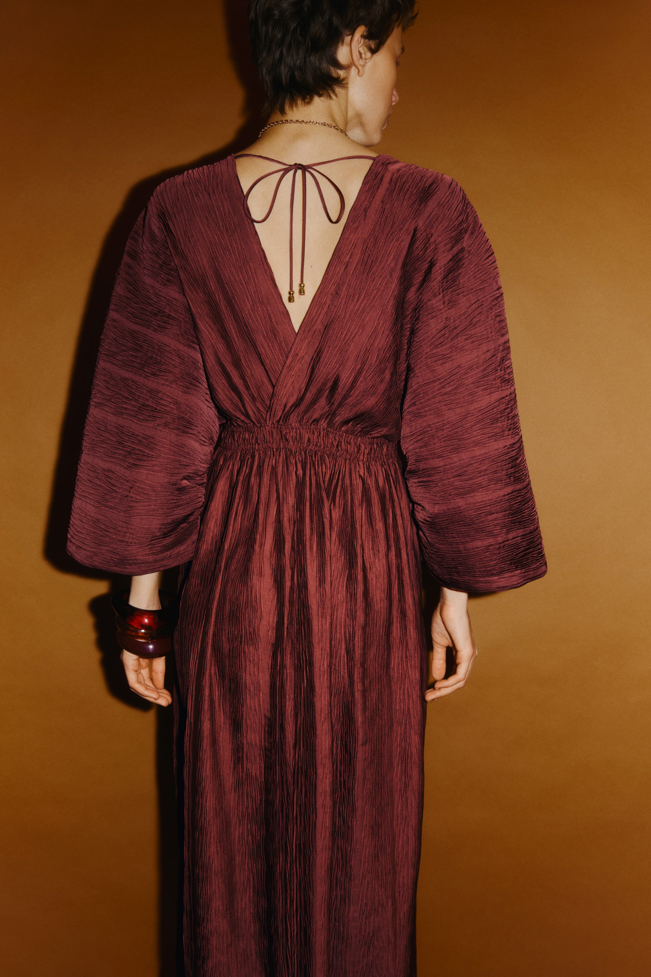 Grösseres Bild ansehen: Ein burgunderfarbenes Maxikleid mit Crinkle-Optik ist von hinten zu sehen. Es hat einen tiefen V‑Ausschnitt mit einem Bindeband, das in goldfarbenen Perlen endet. Das Kleid ist mit weiten, ellbogenlangen Ärmeln und einer betonten Taille gestaltet und fällt in einen bodenlangen Rockteil, alles aus einheitlich strukturiertem Stoff.