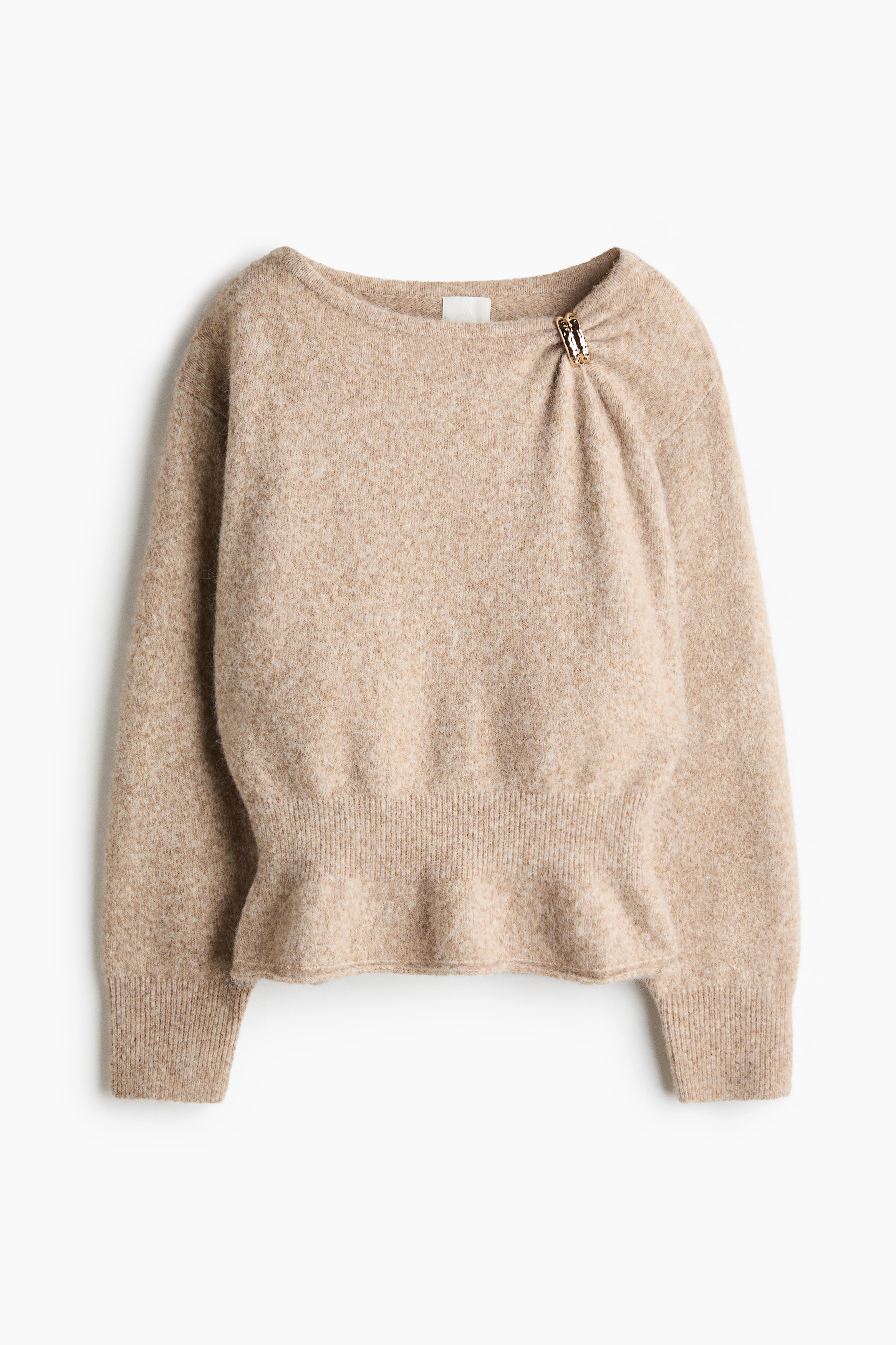 A beige, brushed-knit jumper is being worn by the model with arms crossed, highlighting its boat neckline and dropped shoulders. The jumper features a textured gold-coloured metal detail that cinches the fabric at one side of the collar.