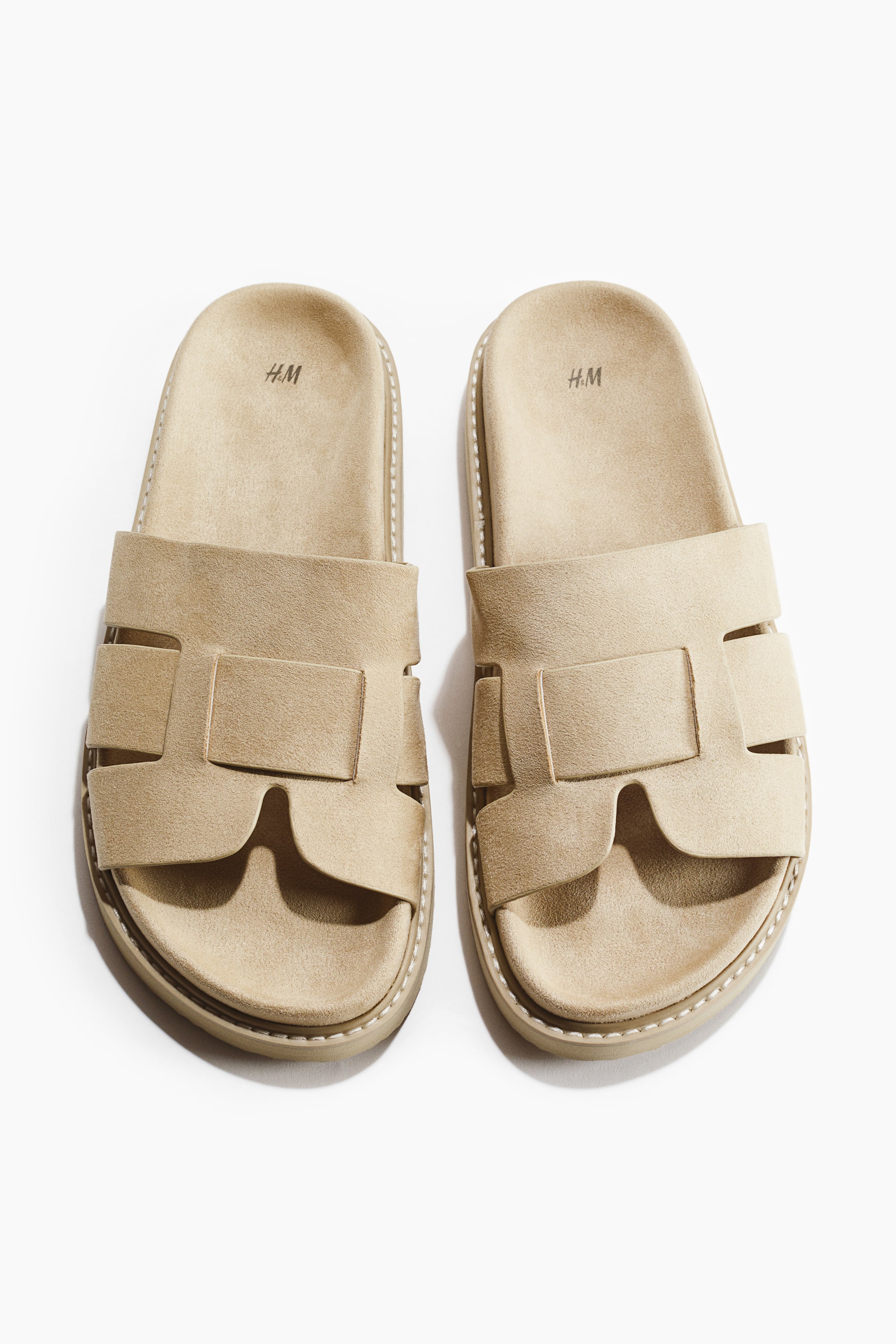 View larger image: A pair of light brown slides showcasing a wide strap with H-shaped cutouts across the forefoot, with the slides oriented frontward. Each slide boasts a thick sole with noticeable white stitching around the edges and "H.M" debossed on the footbed.
