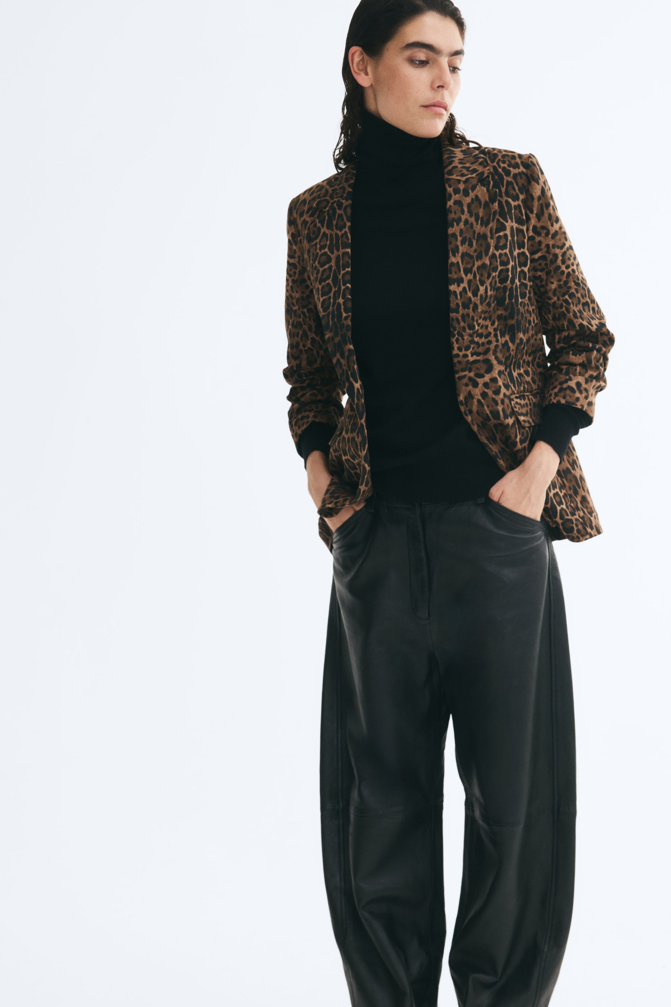 View larger image: A leopard-print fitted blazer is worn open by the model, featuring notch lapels, jetted front pockets with flaps, and no fastenings. The blazer is layered over a black roll-neck top and paired with wide-leg black trousers.
