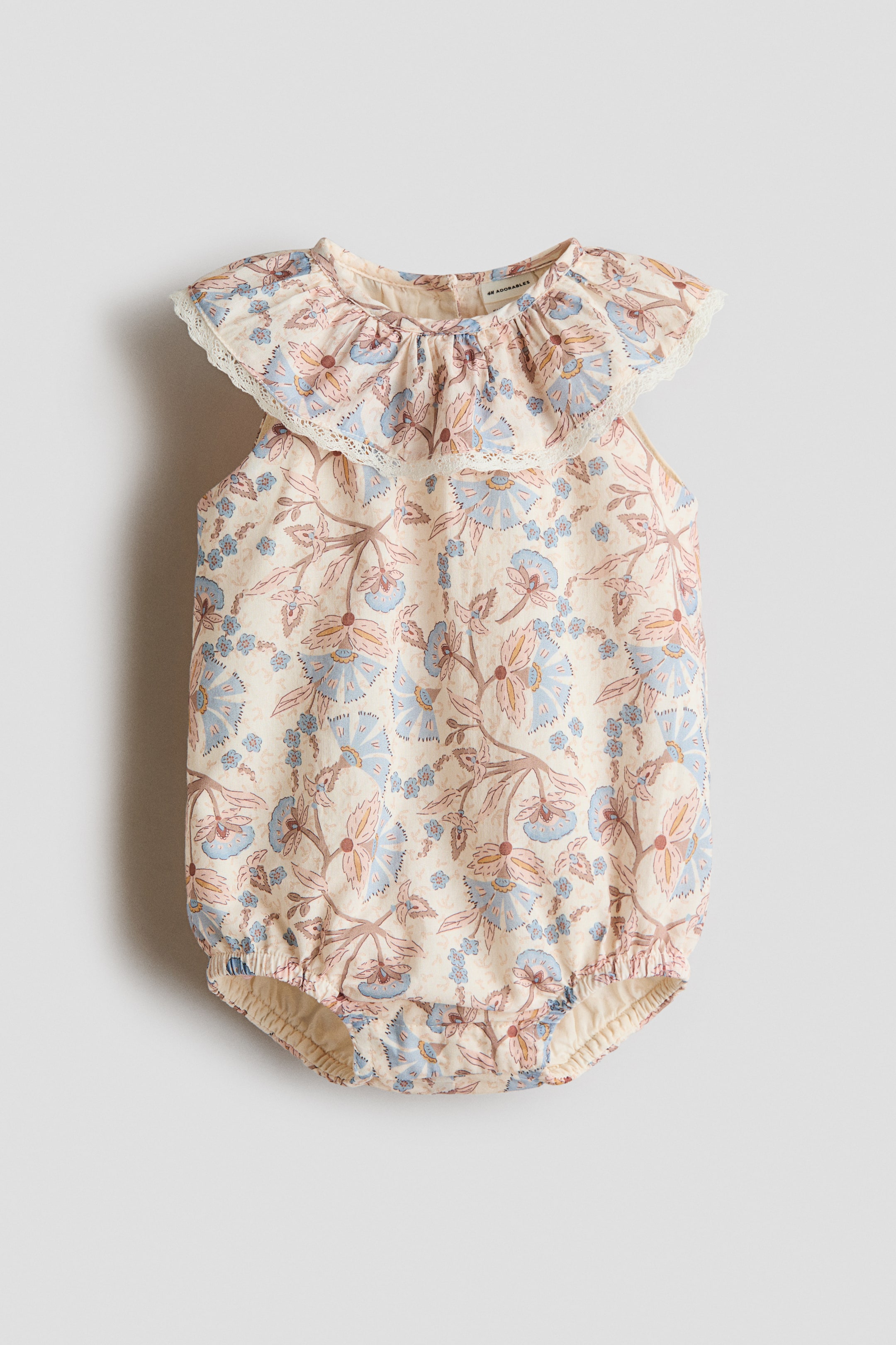 View larger image: A sleeveless romper suit is showcased from the front, crafted from a soft weave fabric adorned with a delicate floral pattern in shades of blue and muted pink against a cream backdrop. It features a ruffled round collar and ruffled armholes, both highlighted with broderie anglaise trim, with elastication visible around the leg openings.