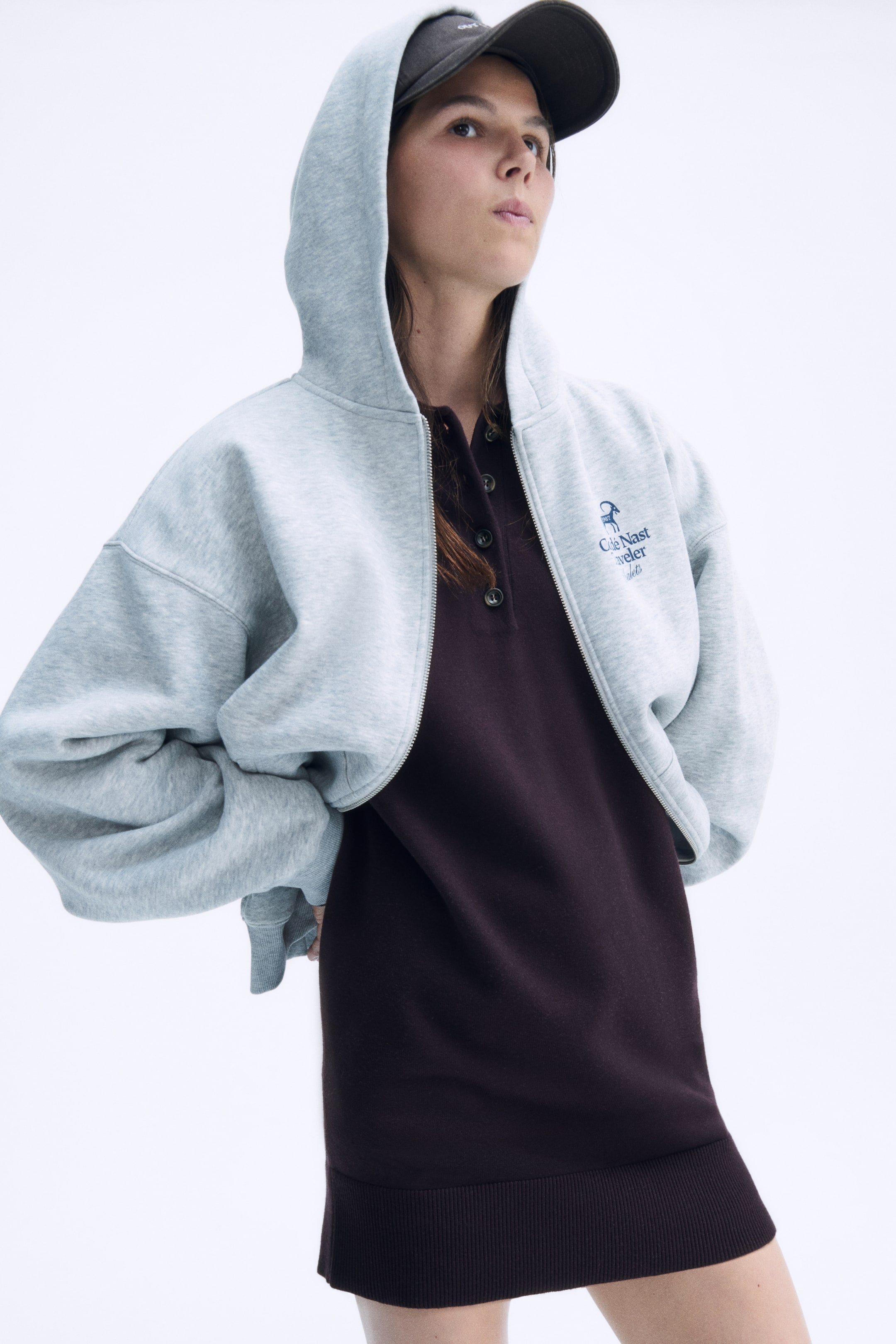 Größeres Bild ansehen: Ein kurzes, dunkelbraunes Feinstrickkleid ist von vorne zu sehen und verfügt über einen Kragen, eine Knopfleiste und einen breiten Rippbund. Das Kleid ist unter einem offenen hellgrauen Hoodie mit Reißverschluss getragen und betont so seine lockere Passform und die weiche Textur.