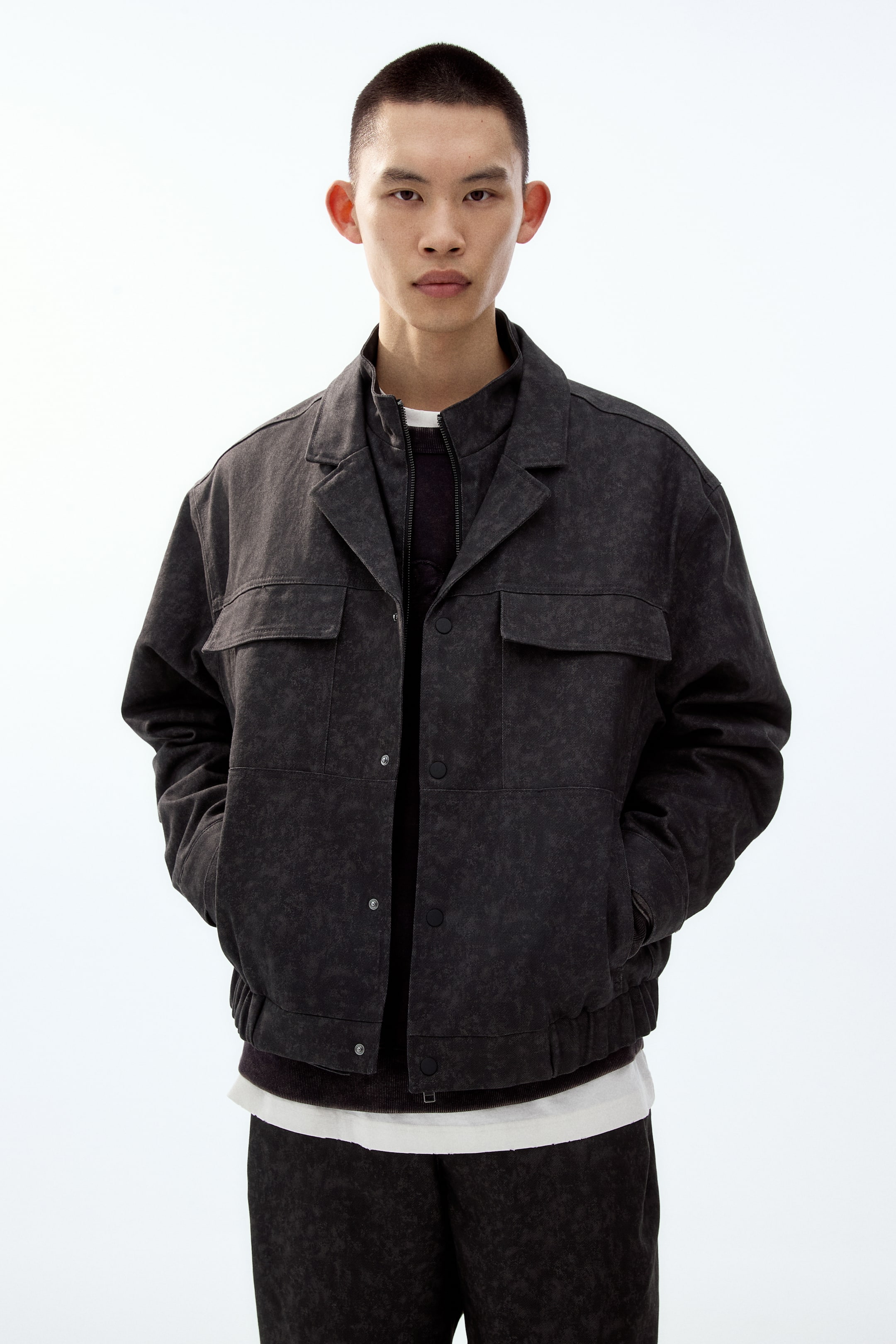 이미지 확대 보기: 정면이 보이는 짙은 회색 재킷으로, 워싱 처리된 질감이 돋보이며 뾰족한 칼라와 버튼 앞여밈이 특징입니다. 가슴 플랩 포켓 2개와 사이드 포켓 2개를 갖추고 있으며 리브드(시보리) 밑단이 있고, 어두운 색 지퍼 스웨터와 흰색 티셔츠 위에 오픈 상태로 레이어드되어 있습니다.