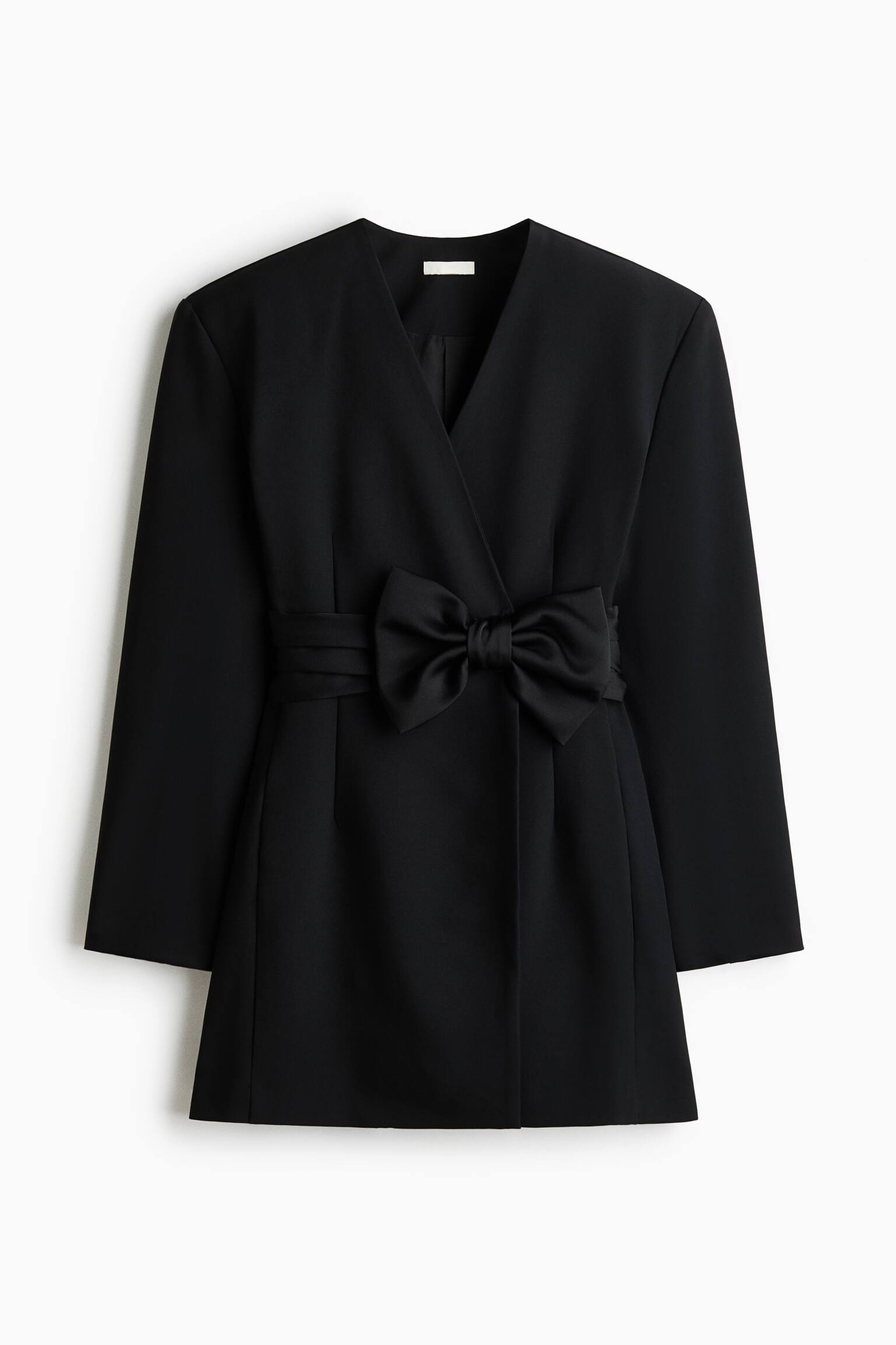 A black blazer dress is shown from the front, featuring a lapelless V-neckline, structured shoulders, and long sleeves. A pleated satin waistband with a large decorative bow accentuates the wrap-front waist.