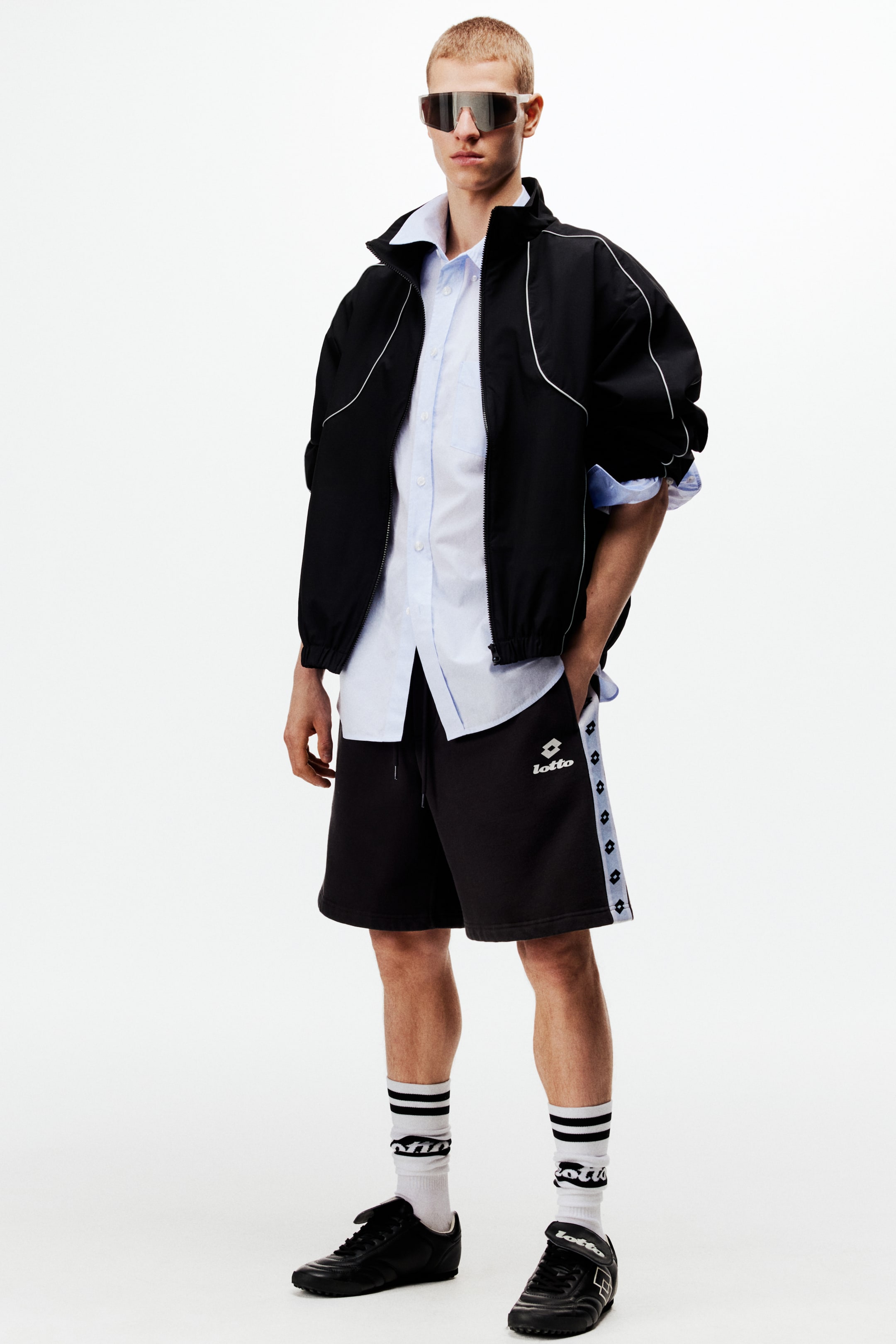 View larger image: A complete sportswear ensemble is being presented, featuring a black track jacket with white piping detail layered over a light blue button-up shirt. The ensemble includes black athletic shorts with a light blue side stripe displaying repeating logos, white crew socks with black stripes, and black athletic shoes, all accessorized with silver mirrored sunglasses.