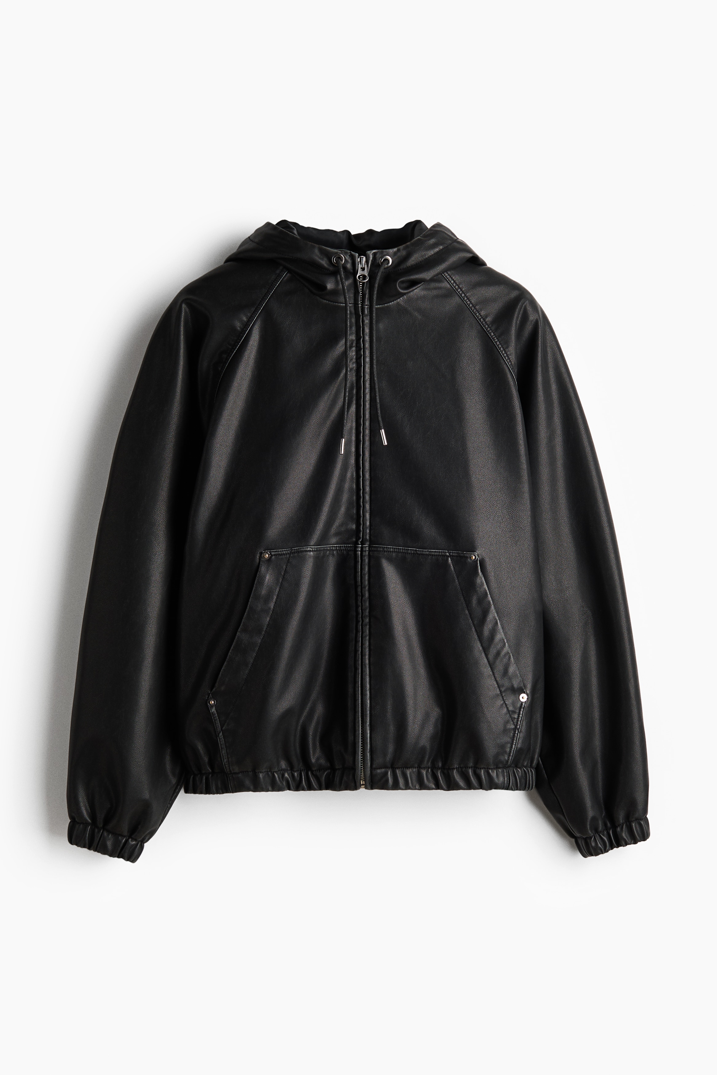 A black jacket in coated fabric featuring a drawstring hood, a front zipper, and elasticated cuffs and hem is worn by the model. The model is layering the jacket over a plaid high-neck shirt and dark jeans while keeping their hands in their jean pockets.