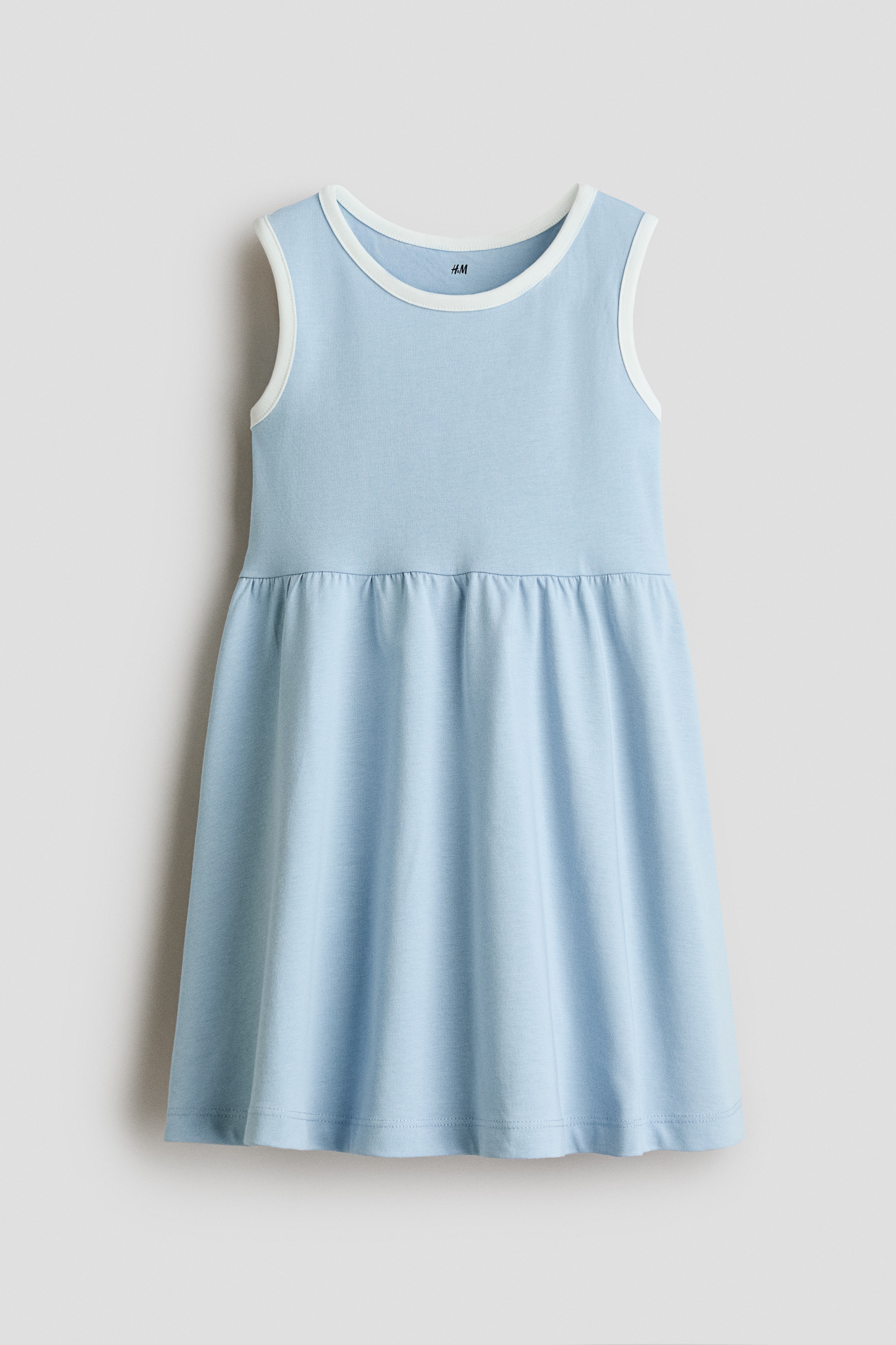 A light blue sleeveless dress with contrasting white trim at the round neckline is worn by the model. She is posing with her chin resting on her hands, highlighting the garmentâs clean, simple design.