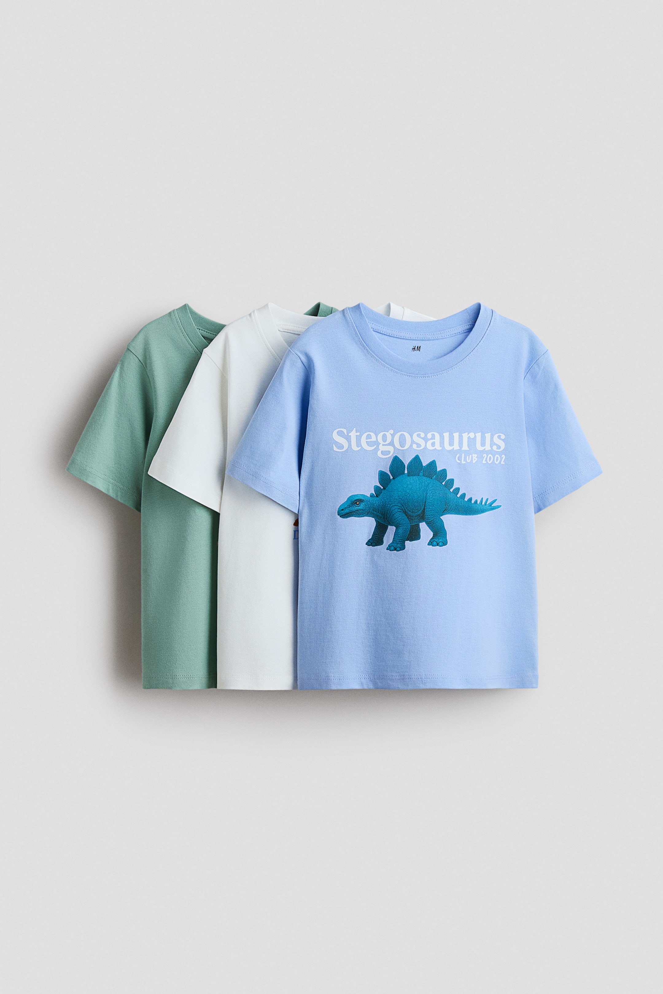 Voir une image plus grande: Trois t-shirts à manches courtes sont superposés : le t-shirt bleu clair à l’avant présente, en vue de face, un imprimé d’un stégosaure bleu et le texte "Stegosaurus CLUB 2002". Un t-shirt blanc cassé est superposé derrière le bleu, et un t-shirt vert sauge est superposé derrière le blanc cassé.