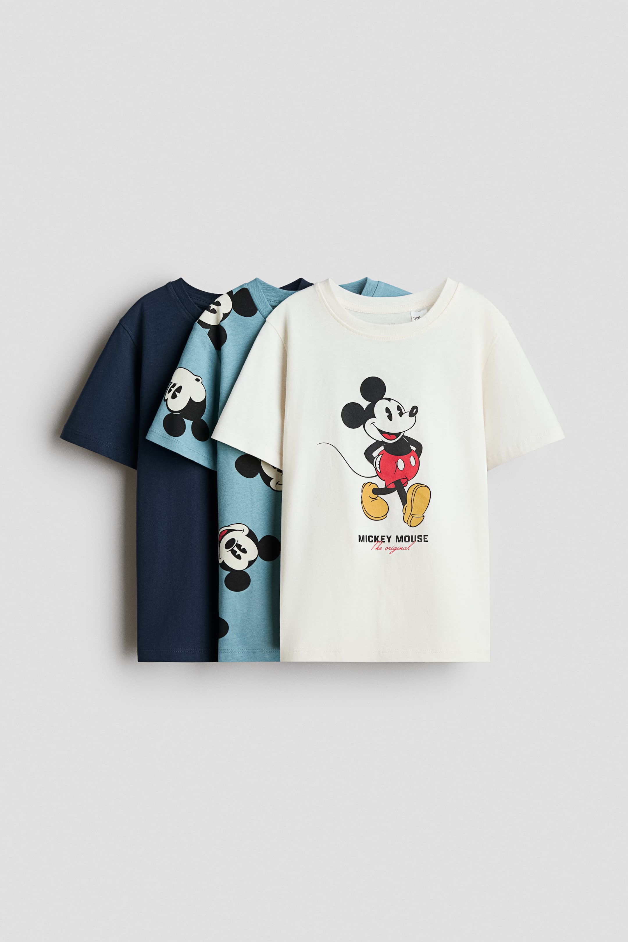 View larger image: Three classic cotton jersey T-shirts with rib-trimmed crew necklines and short sleeves are layered and shown from the front. The front off-white tee features a large central graphic of Mickey Mouse with text below, the middle light-blue tee features an all-over Mickey Mouse pattern, and the back tee is a solid dark navy blue.