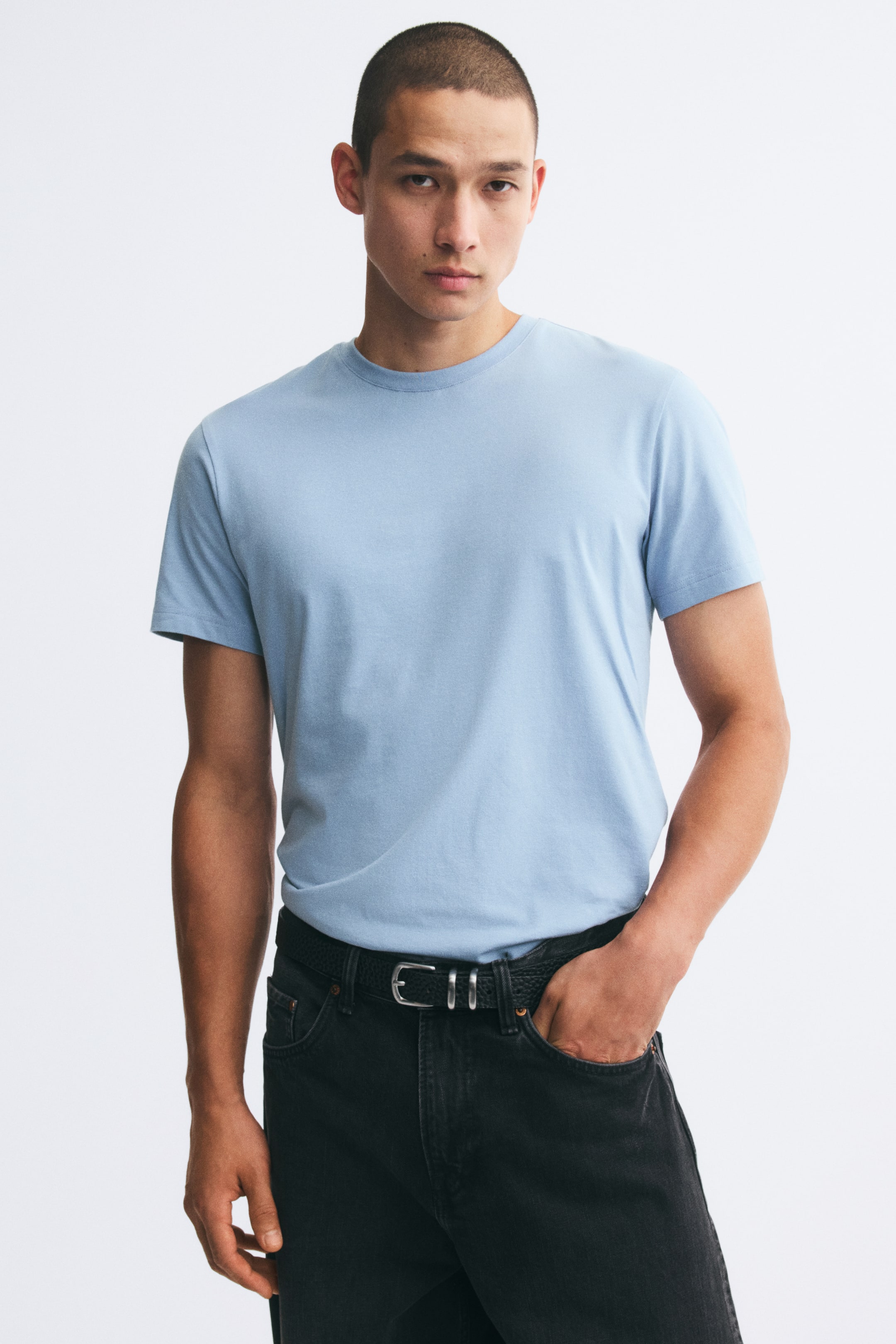 Se større billede: En lyseblå, slim-fit T-shirt i let bomuldsjersey bæres af modellen og er stukket ned i sorte jeans med bælte. T-shirten med korte ærmer har en rund, ribkantet halsudskæring og sidder tæt til kroppen.