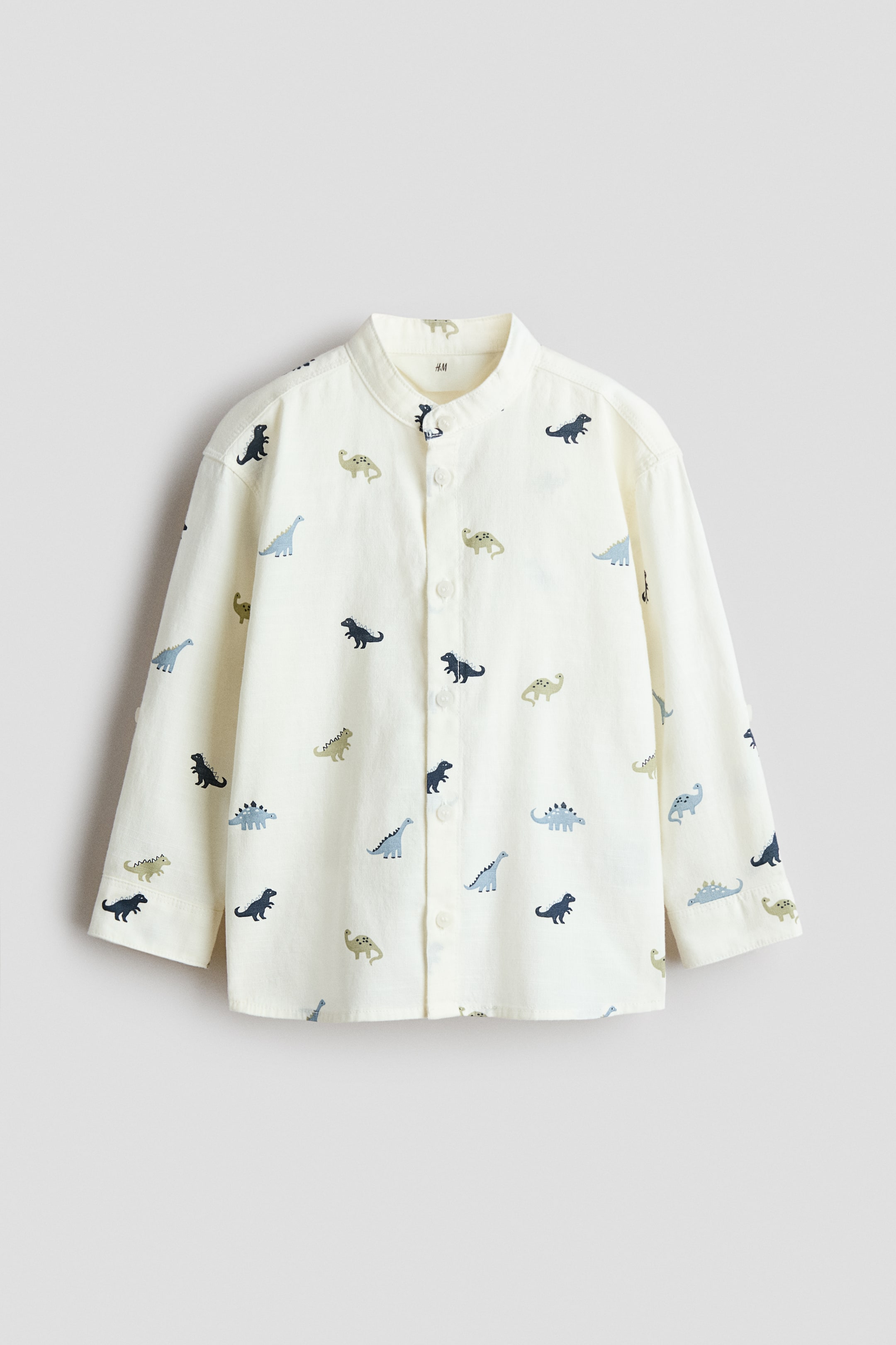 View larger image: A cream shirt, facing FRONT, showcases a band collar with buttons running down the front. This soft, airy cotton weave shirt displays an all-over dinosaur print in dark blue, light blue, and green hues, featuring long sleeves, buttoned cuffs, and a gently rounded hem.