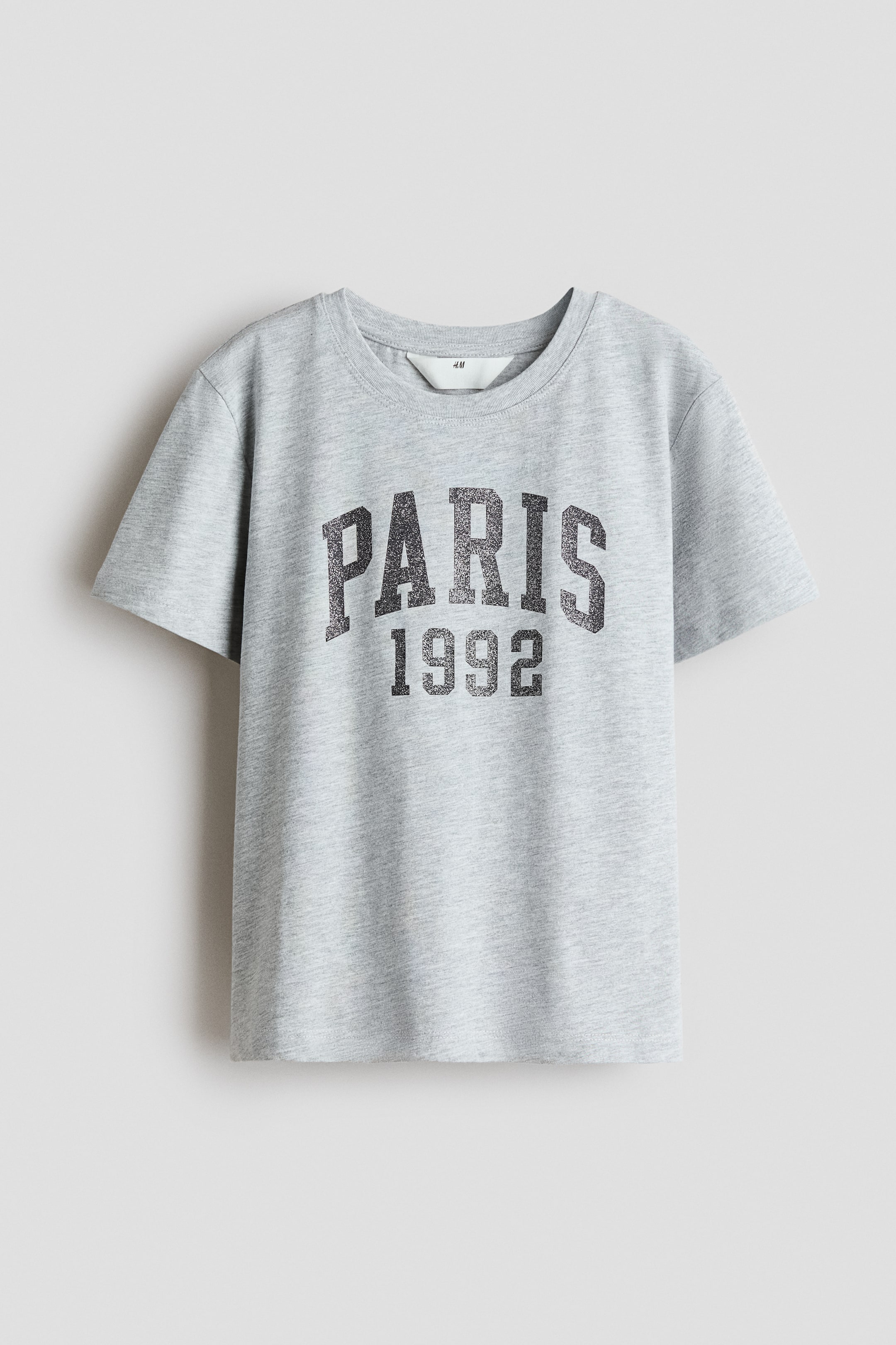 Agrandir l'image: Un T-shirt gris chiné présente sa face avant, arborant un motif imprimé gris foncé vieilli avec "PARIS" en arc au-dessus de "1992". Une bordure côtelée souligne l'encolure ras-du-cou.