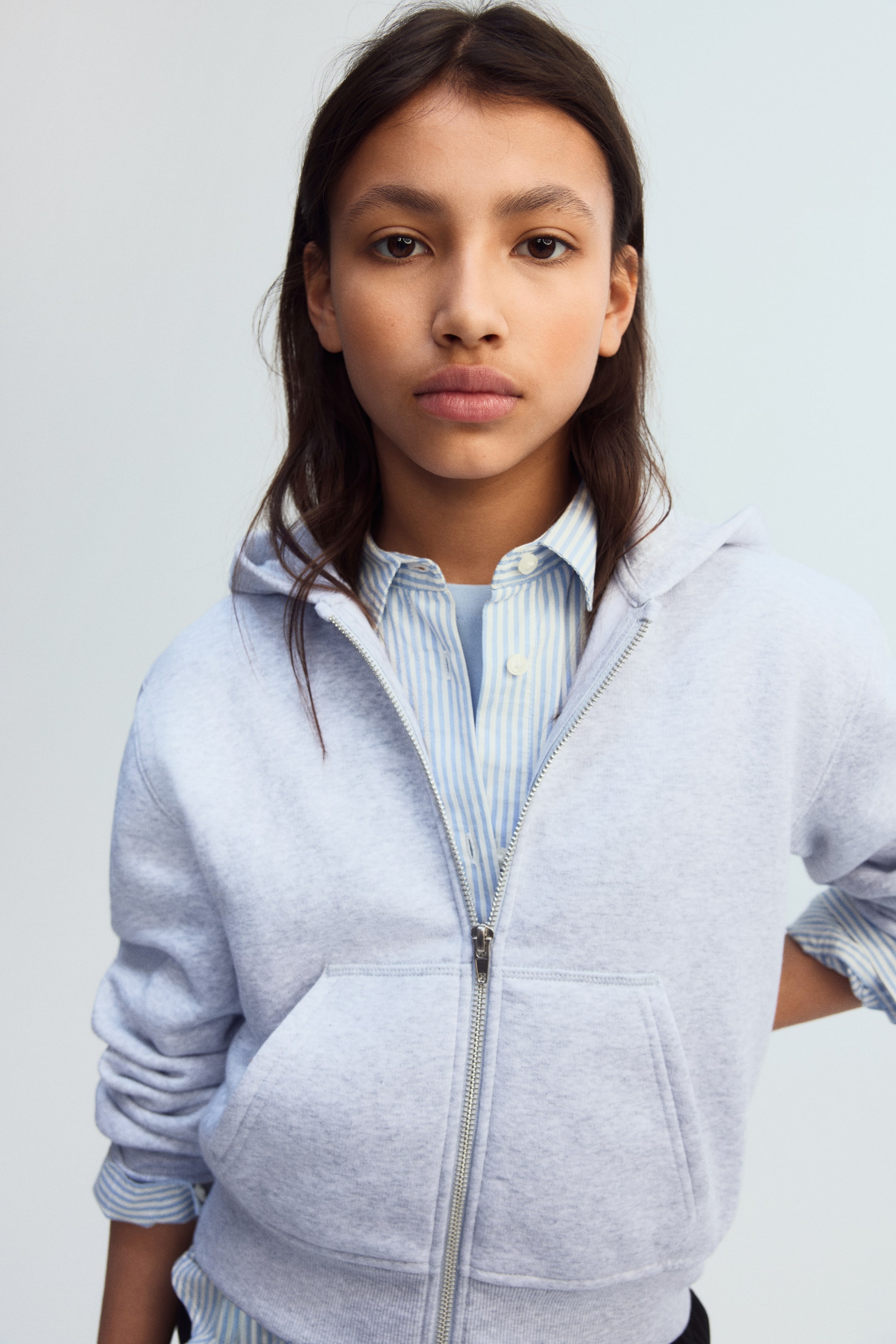 Agrandir l'image: Un hoodie gris clair à fermeture éclair complète est présenté de face, avec une fermeture éclair argentée partiellement ouverte, une poche kangourou à l'avant et des poignets et un ourlet côtelés. Il est superposé à une chemise à boutons rayée bleu clair et blanche.