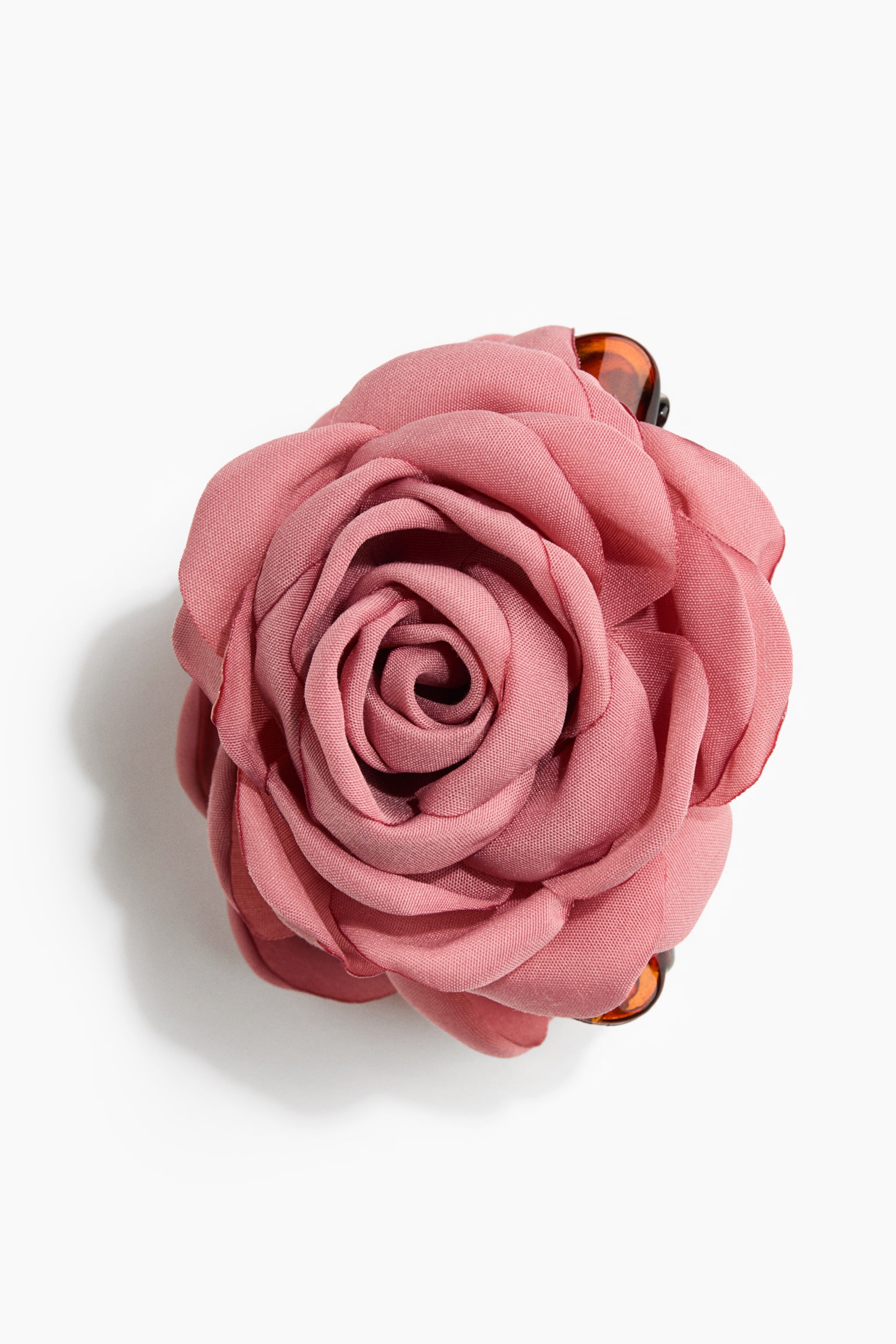 Une pince Ã  cheveux en forme de rose en tissu rose poudrÃ© est prÃ©sentÃ©e de face, mettant en valeur une grande fleur Ã  plusieurs couches aux pÃ©tales texturÃ©s. La rose en tissu est fixÃ©e Ã  une pince Ã  griffes effet Ã©caille de tortue, partiellement visible en haut et en bas.