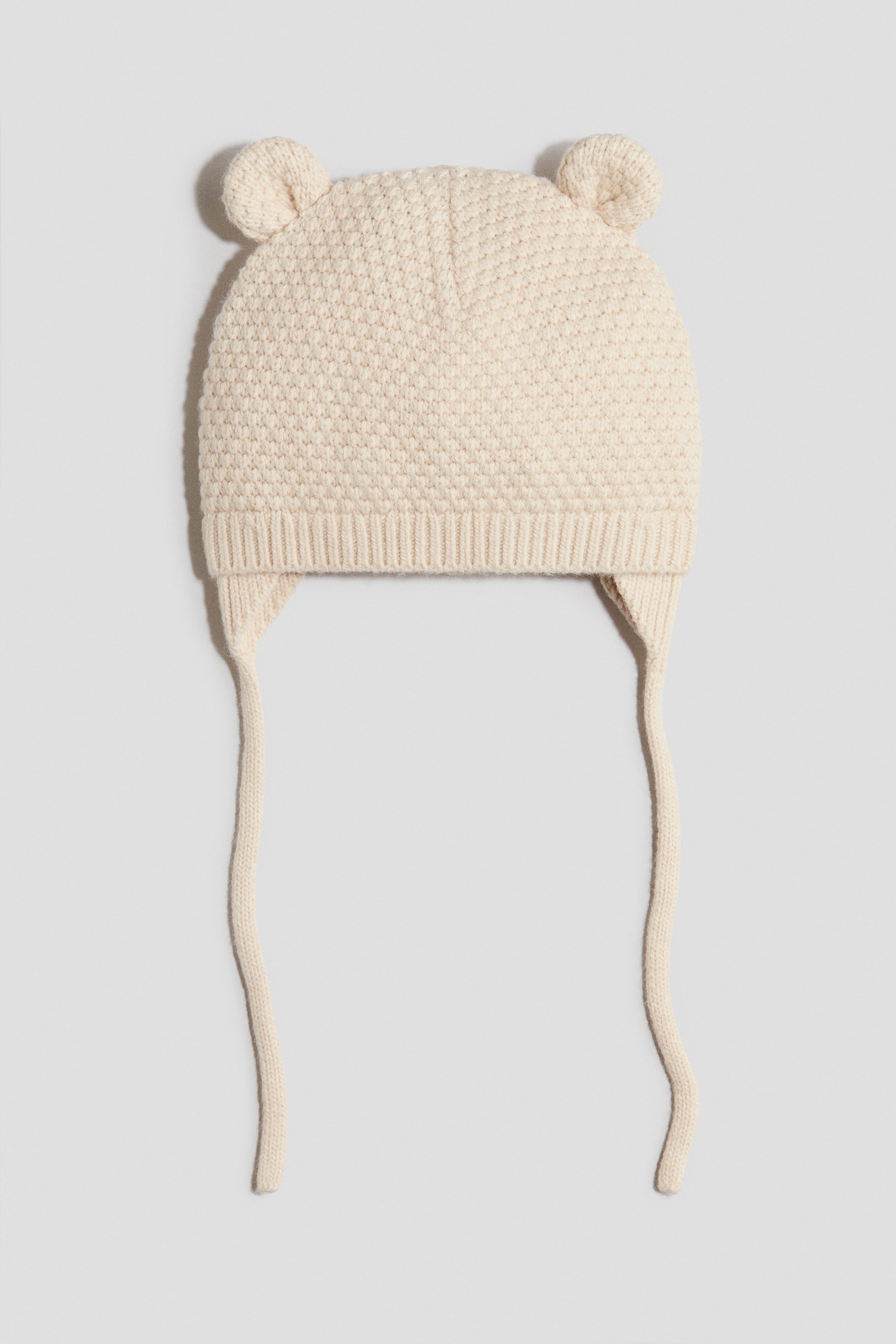 View larger image: A cream-coloured knitted hat is shown from the front, featuring a textured knit pattern on the main body and a ribbed knit band. Two small, rounded ear-like details are positioned on top, with ear flaps extending down from the sides, each with a knitted tie hanging from the end.