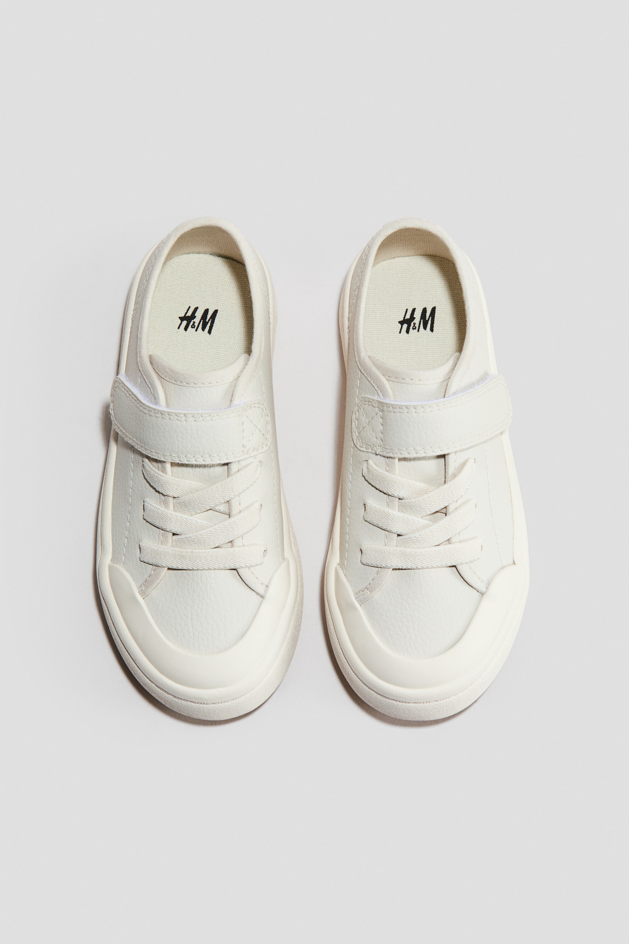 Visa större bild: Två vita sneakers visar en frontvy med texturerade överdelar med dekorativa skosnören och ett enda kardborreband. De har också runda tåhättor och tjocka, något upphöjda sulor.