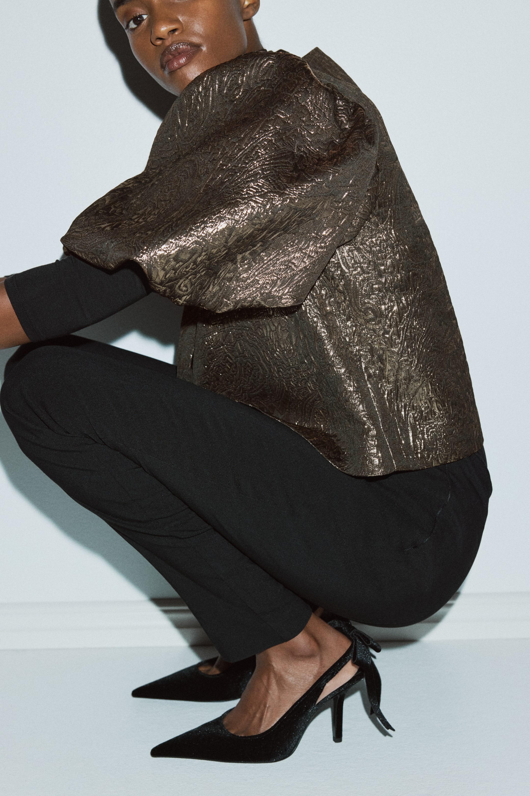 Zobacz większy obraz: Krótka bluzka w solidnym, metalicznym, teksturowanym splocie z obszernymi rękawami balonowymi jest noszona przez modelkę. Jest nałożona na czarną bluzkę z długim rękawem i zestawiona z czarnymi spodniami, podczas gdy modelka klęczy.