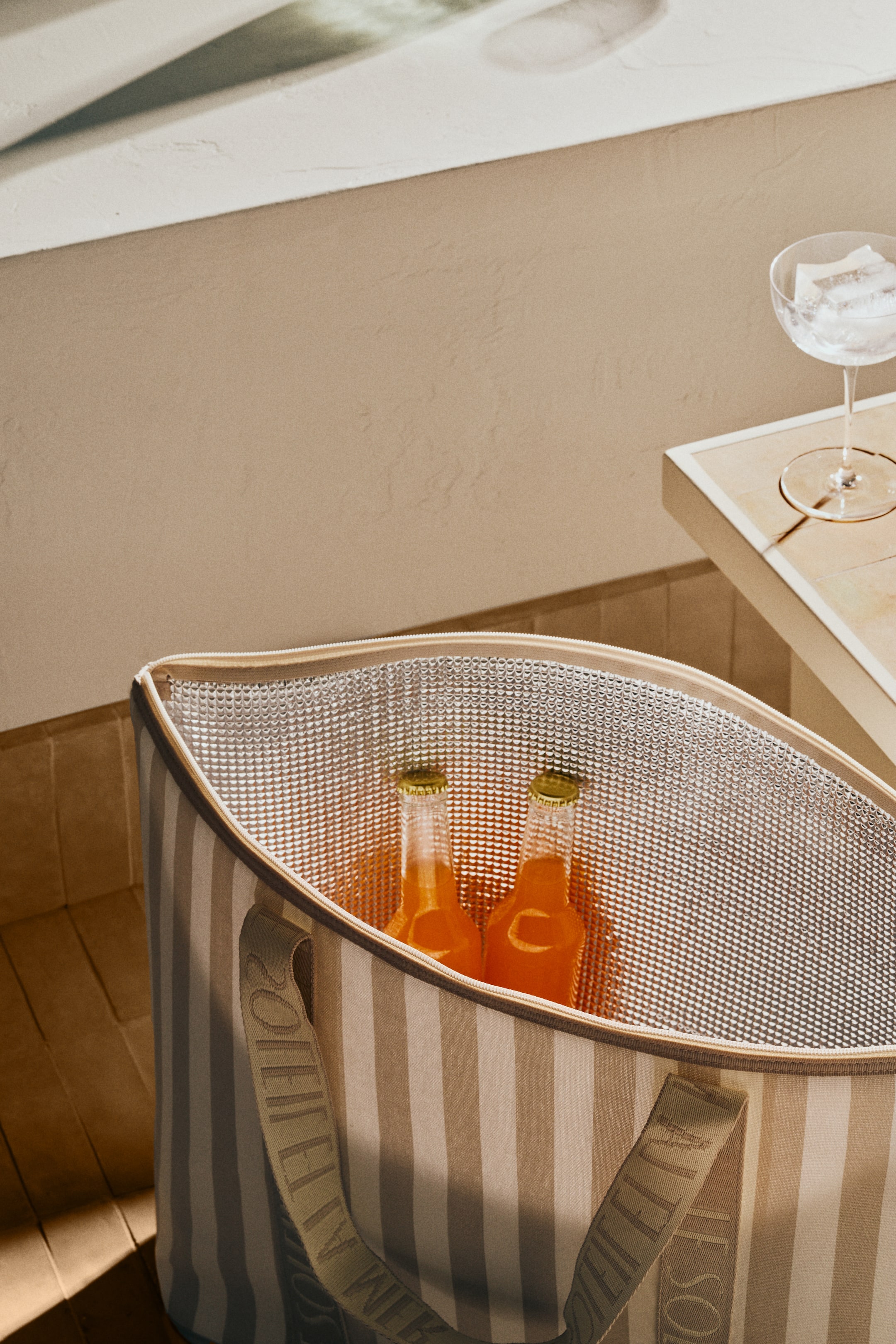 View larger image: The cooler bag is slanted downwards and slightly to the right, showcasing its open top and vertical beige and cream stripes. Its silver insulated lining is visible inside, containing two bottles of orange liquid, while one fabric handle with repeating "SOLEIL" text extends from the side.