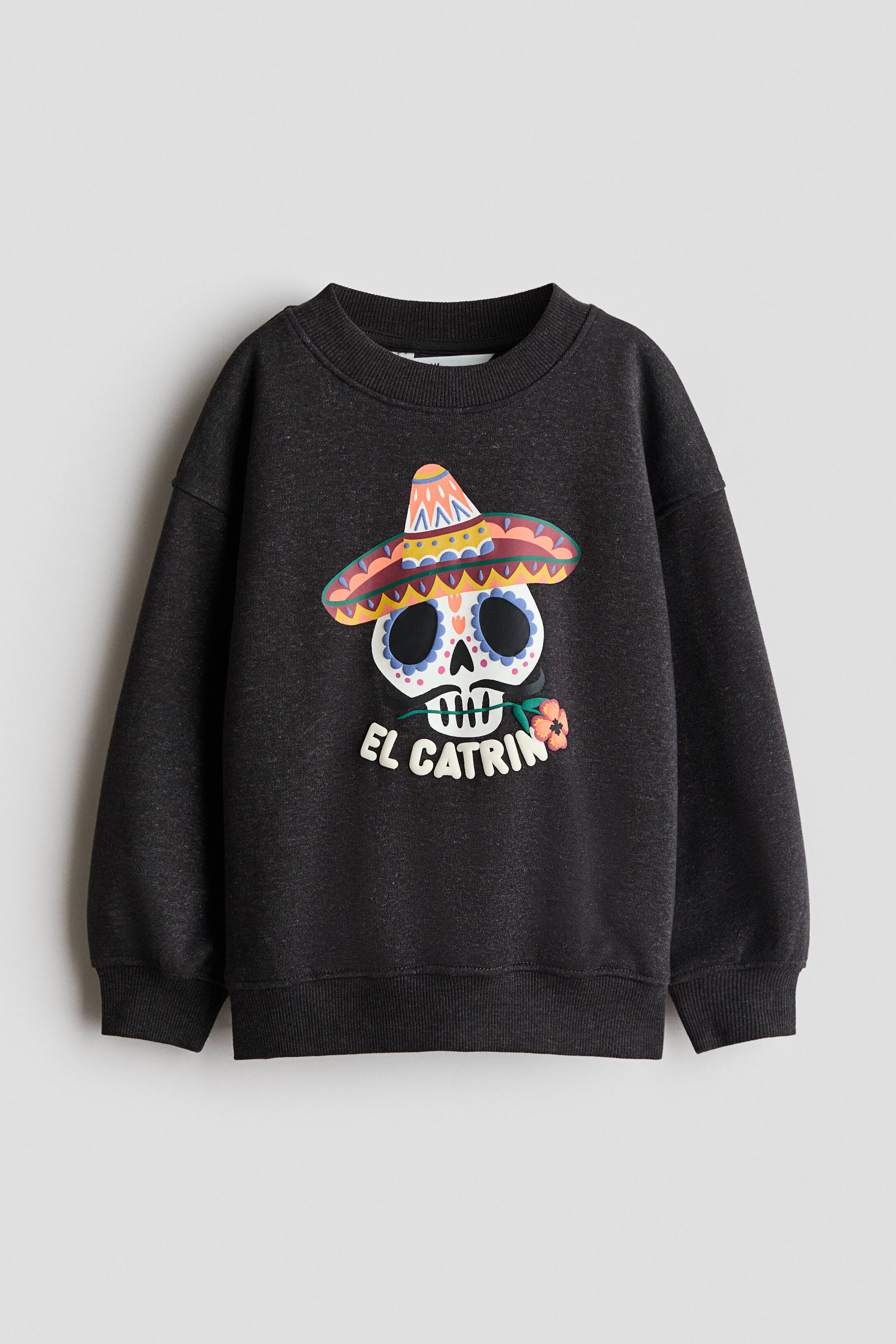 View larger image: A dark gray sweatshirt is facing forward, featuring a crew neck, ribbed cuffs, and a ribbed hem. The front showcases a vibrant graphic of a stylized skull wearing a multi-colored sombrero, holding a pink flower, with the words "EL CATRIN" printed below it.