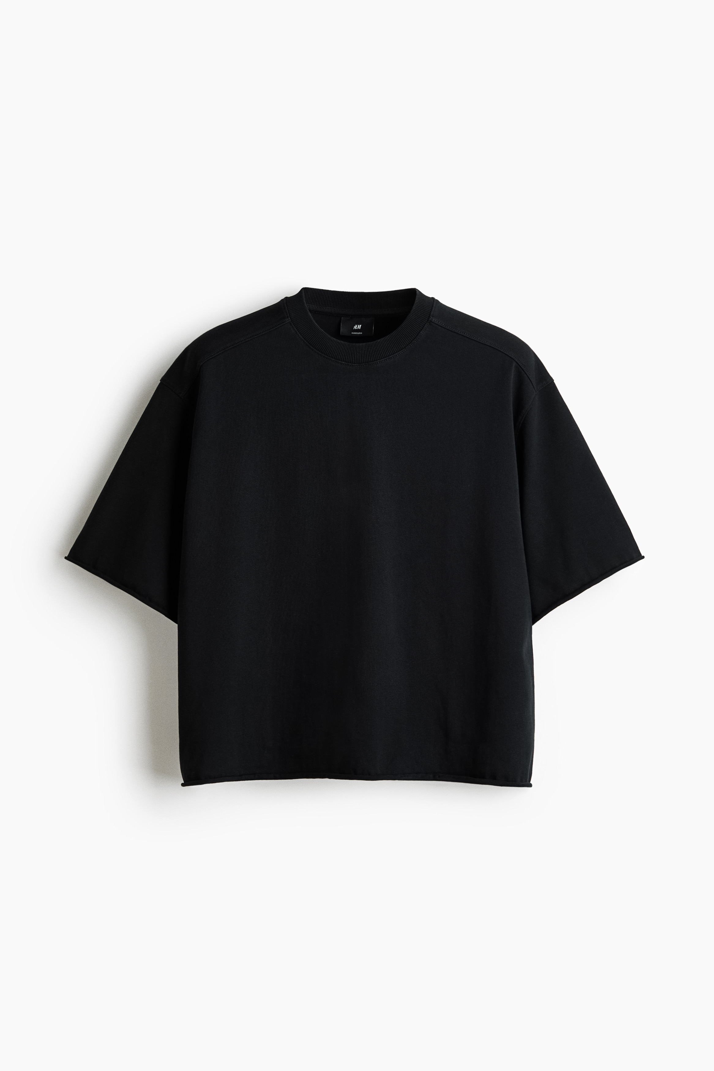 A black, boxy, oversized T-shirt with dropped shoulders and a raw-cut hem is being worn by the model. The shirt features a large, distressed white graphic print across the back and hangs loosely over blue denim jeans.