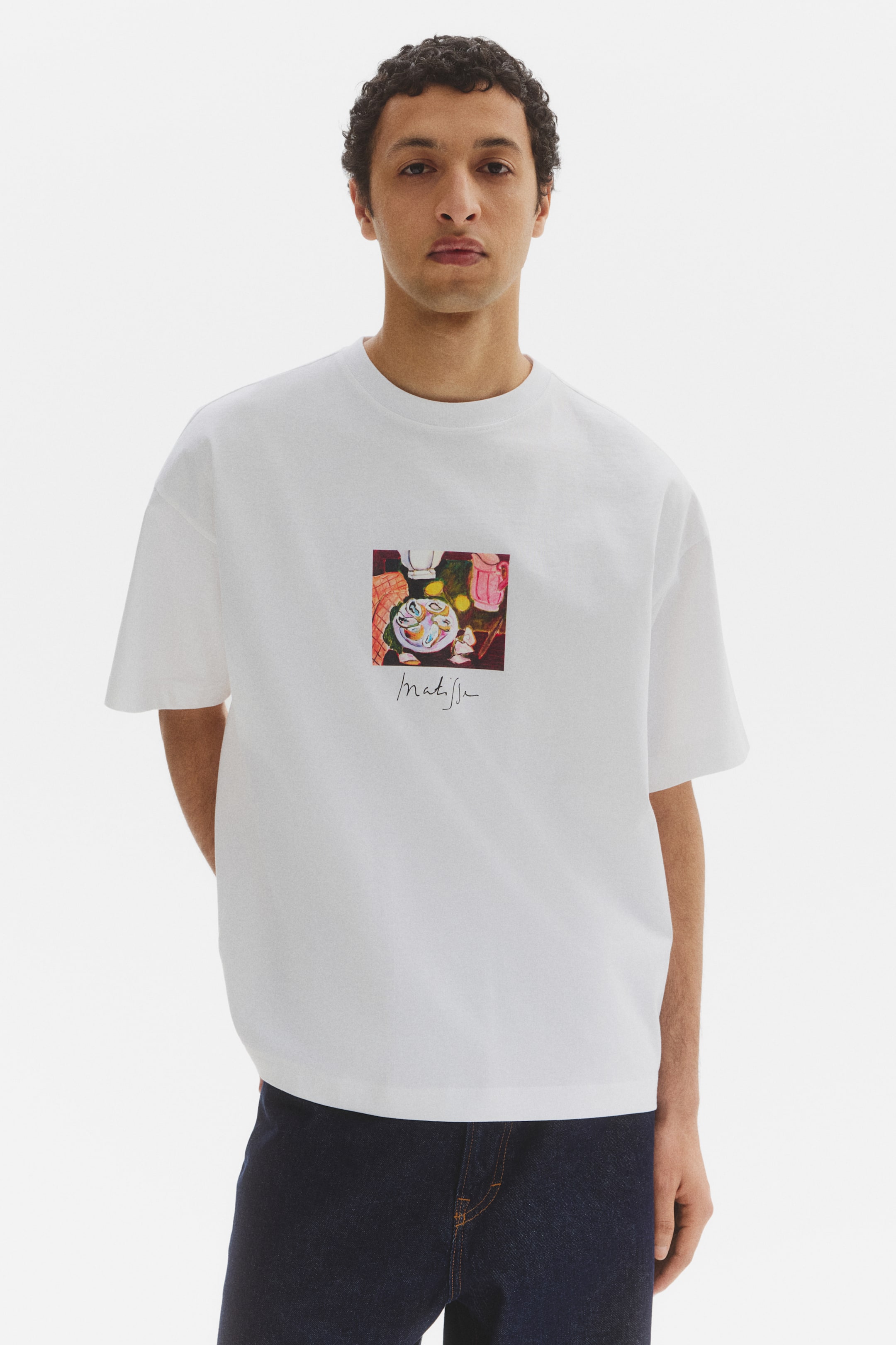 Grotere afbeelding bekijken: Een wit t-shirt met een ronde hals en korte mouwen wordt van voren gepresenteerd en toont een ontspannen pasvorm. Een kleurrijke vierkante grafische print verschijnt op de borst, met daarop een stilleven schilderij en daaronder de handtekening "Matisse".