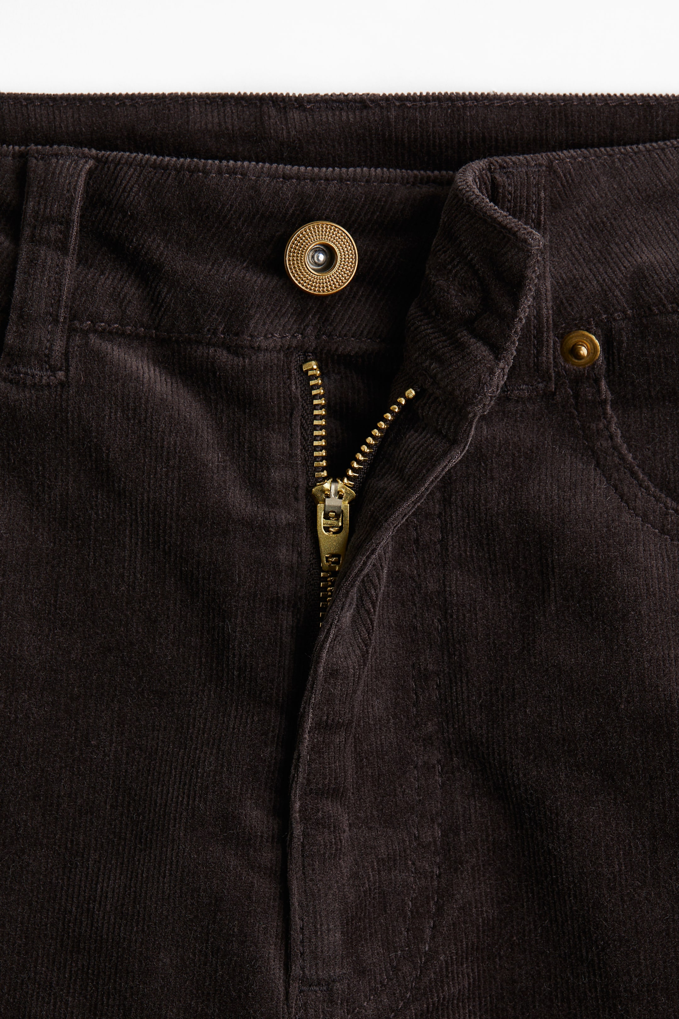 Nagyobb kép megtekintése: Elölnézet sötét kordbársony nadrágról. A ruhadarab magas derékkal rendelkezik, rézszínű gombzárral és részben lehúzott rézszínű cipzárral, valamint látható varrásokkal, amelyek a jobb oldalon lévő elülső zsebnyílást körvonalazzák.