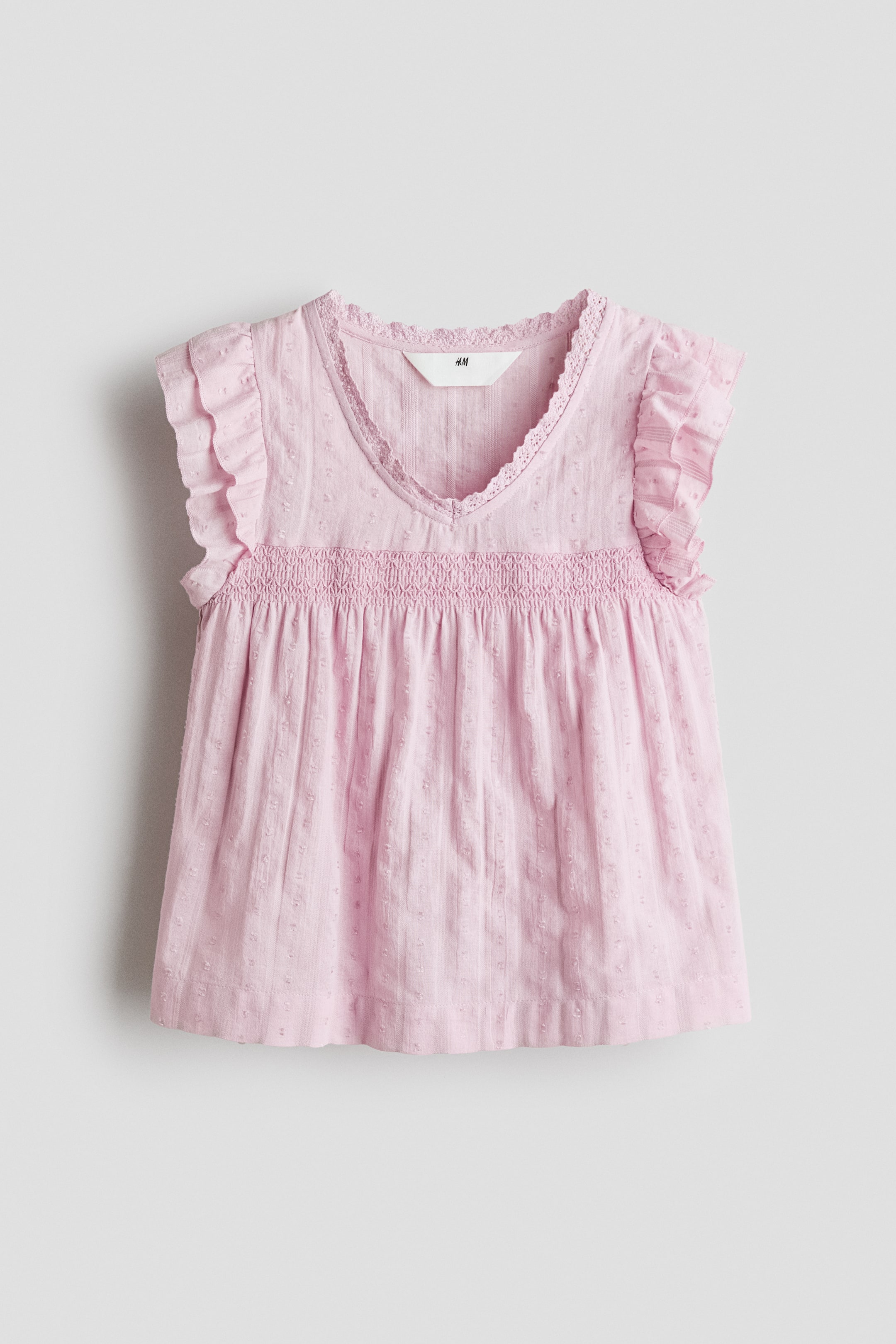 View larger image: The front view of a pink blouse is showcased, highlighting a lace-trimmed V-shaped neckline and double-layered flutter sleeves. The front is adorned with smocking, while the lower part of the blouse displays a subtle dotted texture, ending with a gentle flare at the hem.
