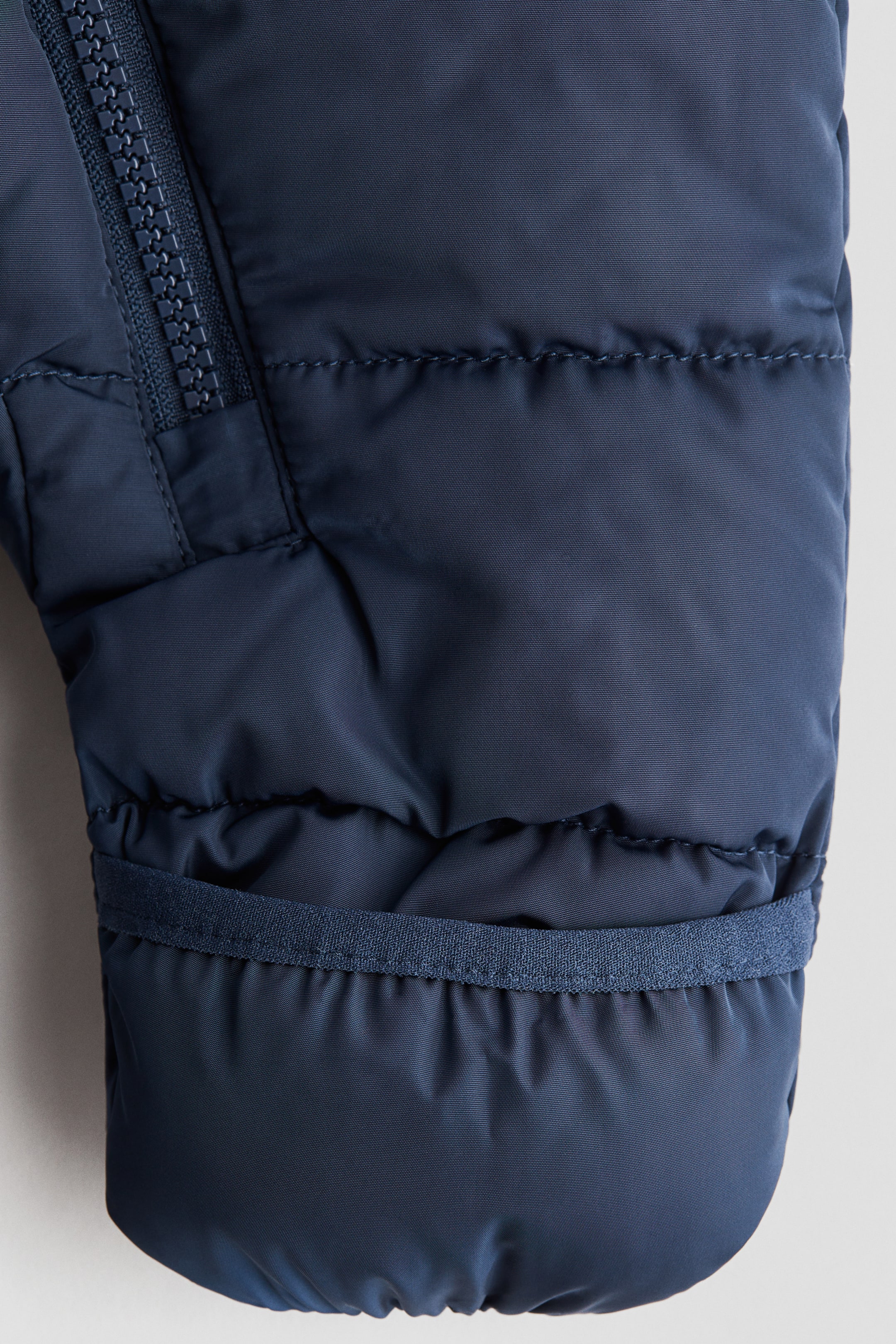 View larger image: A dark blue pile-lined snowsuit is featuring a close-up of its quilted, padded outer shell, with visible stitching creating horizontal sections. A heavy-duty zipper is running diagonally on the upper left, and a flat, elasticized band is appearing horizontally across the lower section, indicating a gathered cuff.
