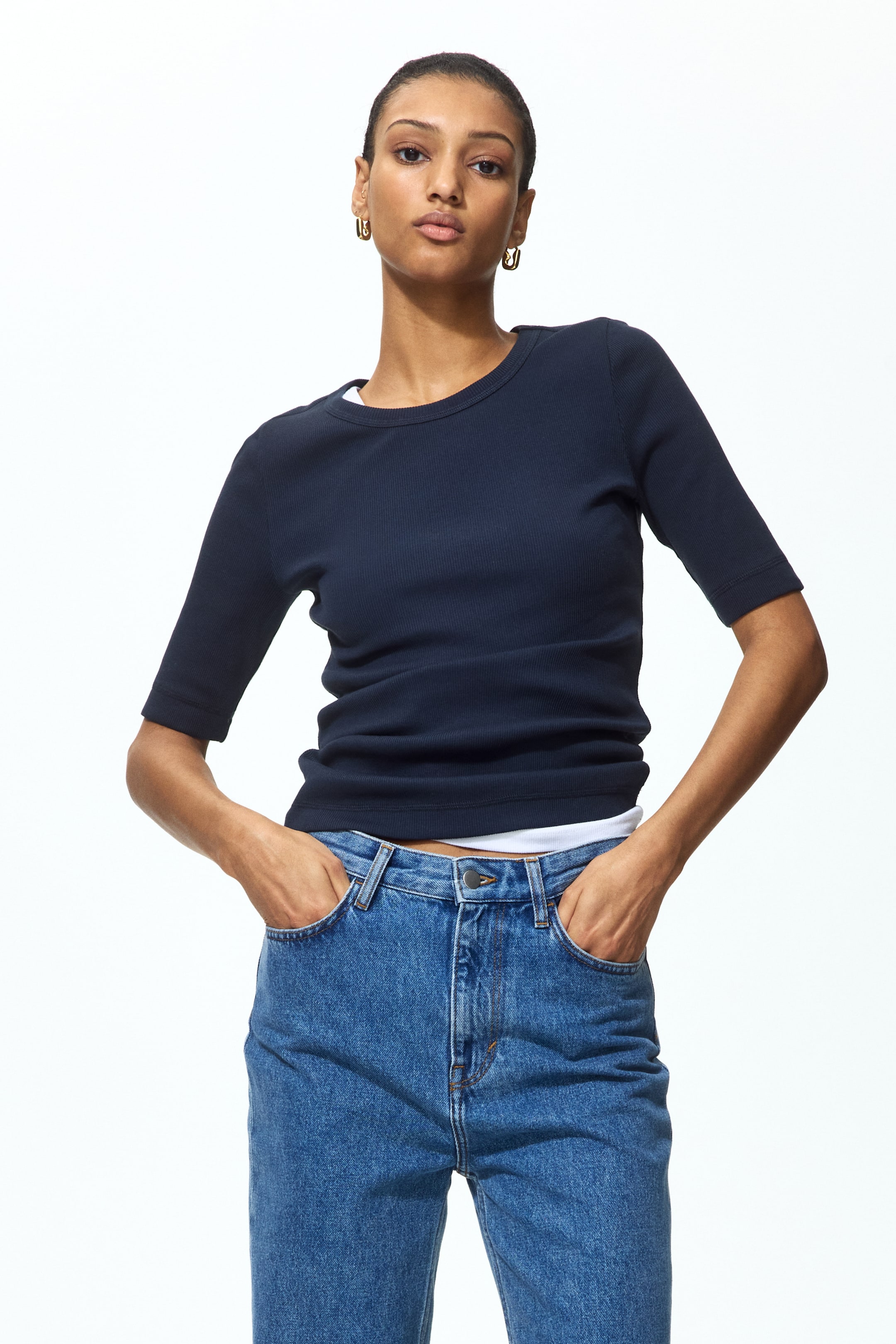 拡大画像を表示: ネイビーブルーのリブトップが着用されており、ラウンドネックと肘丈の袖が特徴です。フィットしたシルエットを呈し、ネックラインと裾から白いアンダーシャツがさりげなく覗いています。