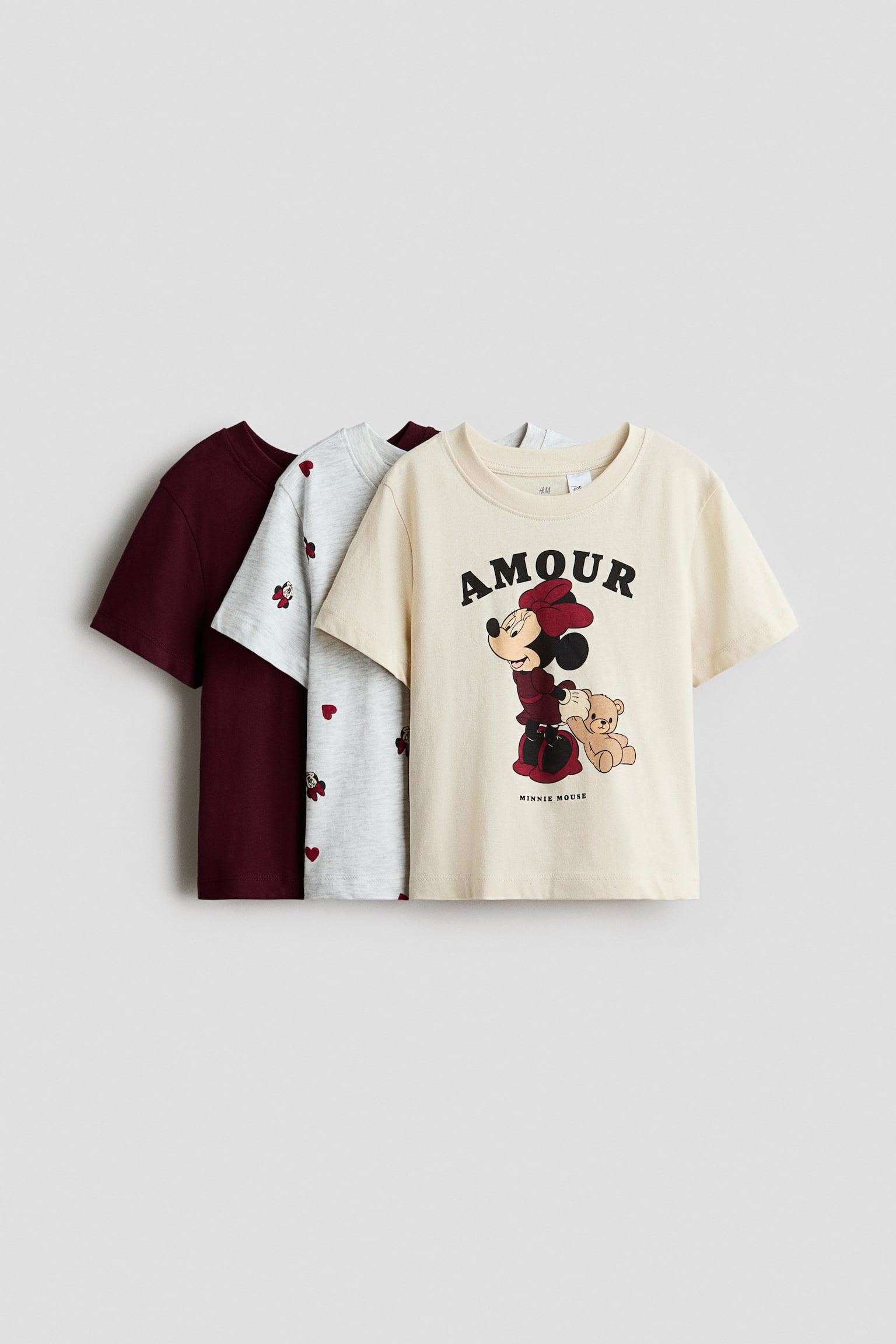 Three cotton jersey t-shirts are shown from the front in an overlapping arrangement. The front cream-coloured t-shirt features a round, rib-trimmed neckline and a graphic print of Minnie Mouse holding a teddy bear with the word "AMOUR" printed above. Layered behind are a light grey t-shirt featuring an all-over Minnie Mouse and heart pattern, and a solid dark burgundy t-shirt.