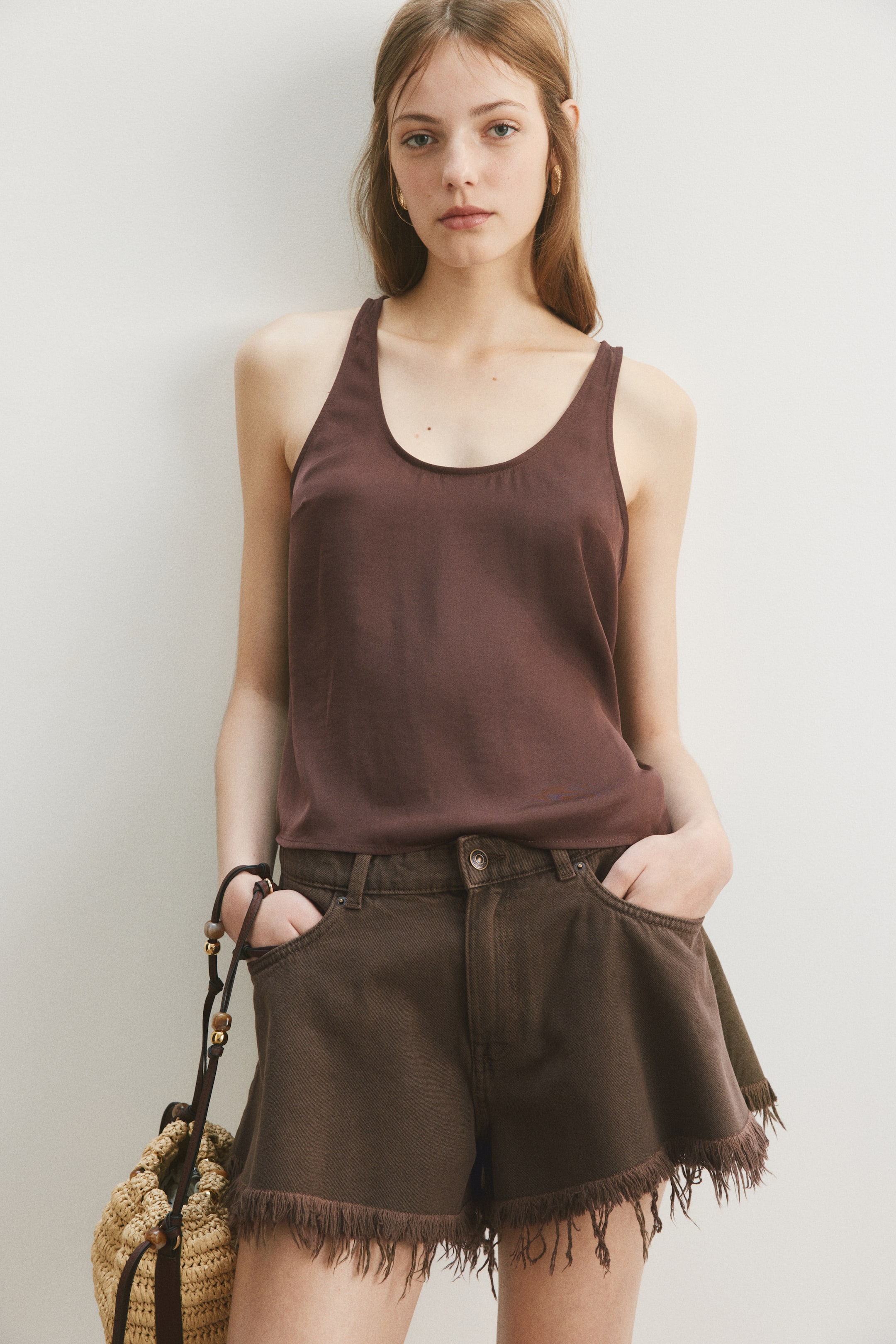 View larger image: The model is wearing a brown sleeveless top crafted from airy chiffon, featuring a scoop neckline and a gently rounded hem. She styles it with brown shorts that have a frayed hem and carries a straw bag.