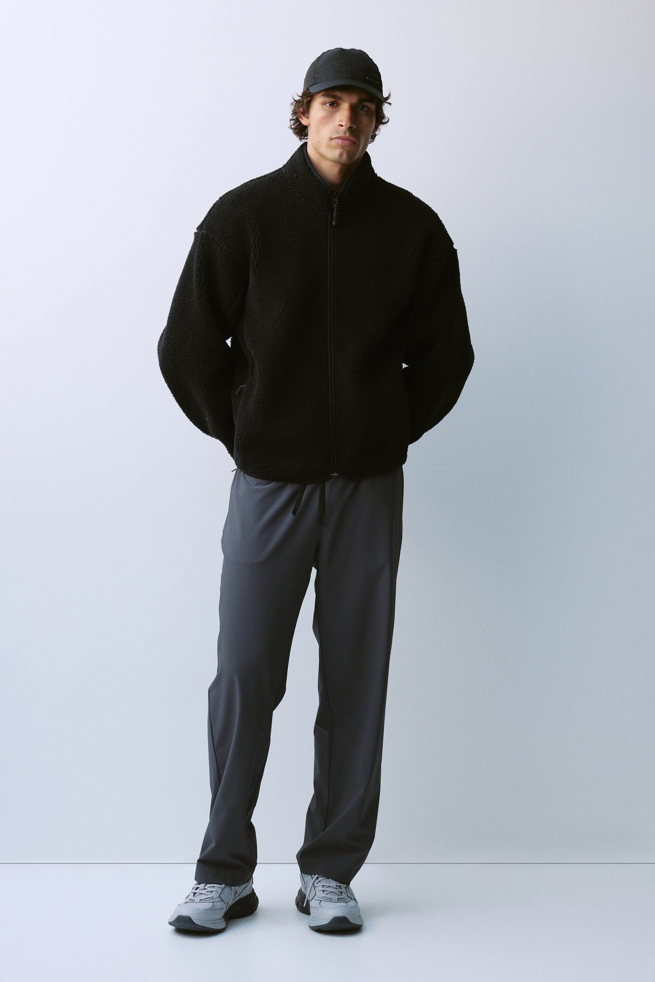 View larger image: A black textured fleece jacket with a high collar and zippered front is being worn by the model. The jacket is styled with dark grey drawstring trousers, a black cap, and grey sneakers.