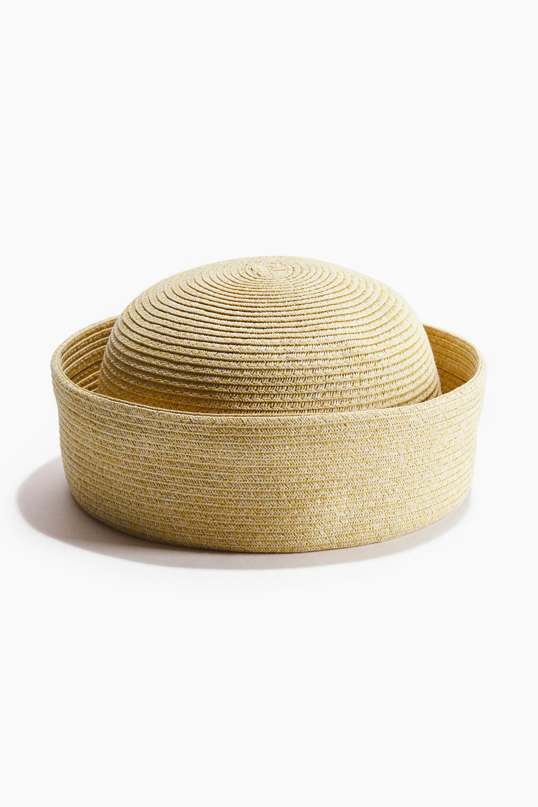 Ver imagen más grande: Un sombrero de paja tejida de color amarillo claro aparece desde un ligero ángulo. El sombrero presenta una corona redonda de cima plana con un tejido concéntrico, que se anida dentro de un ala circular más ancha y plana que exhibe un tejido horizontal.
