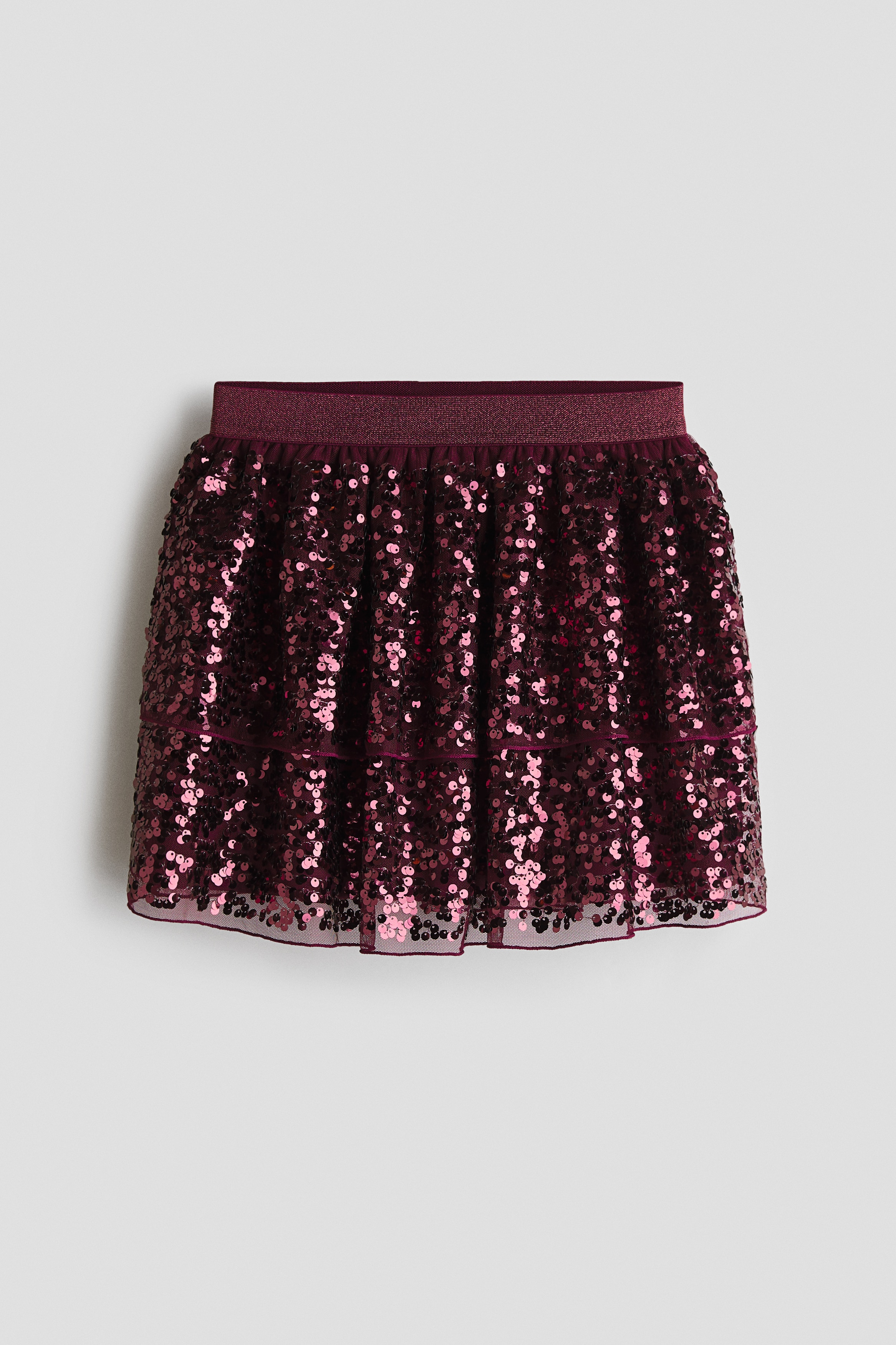 A burgundy tulle skirt covered in shimmering sequins is shown from the front, featuring a tiered, flared design. The skirt is paired with a matching textured sleeveless top, highlighting its glittery elasticated waistband and lightweight, layered structure.