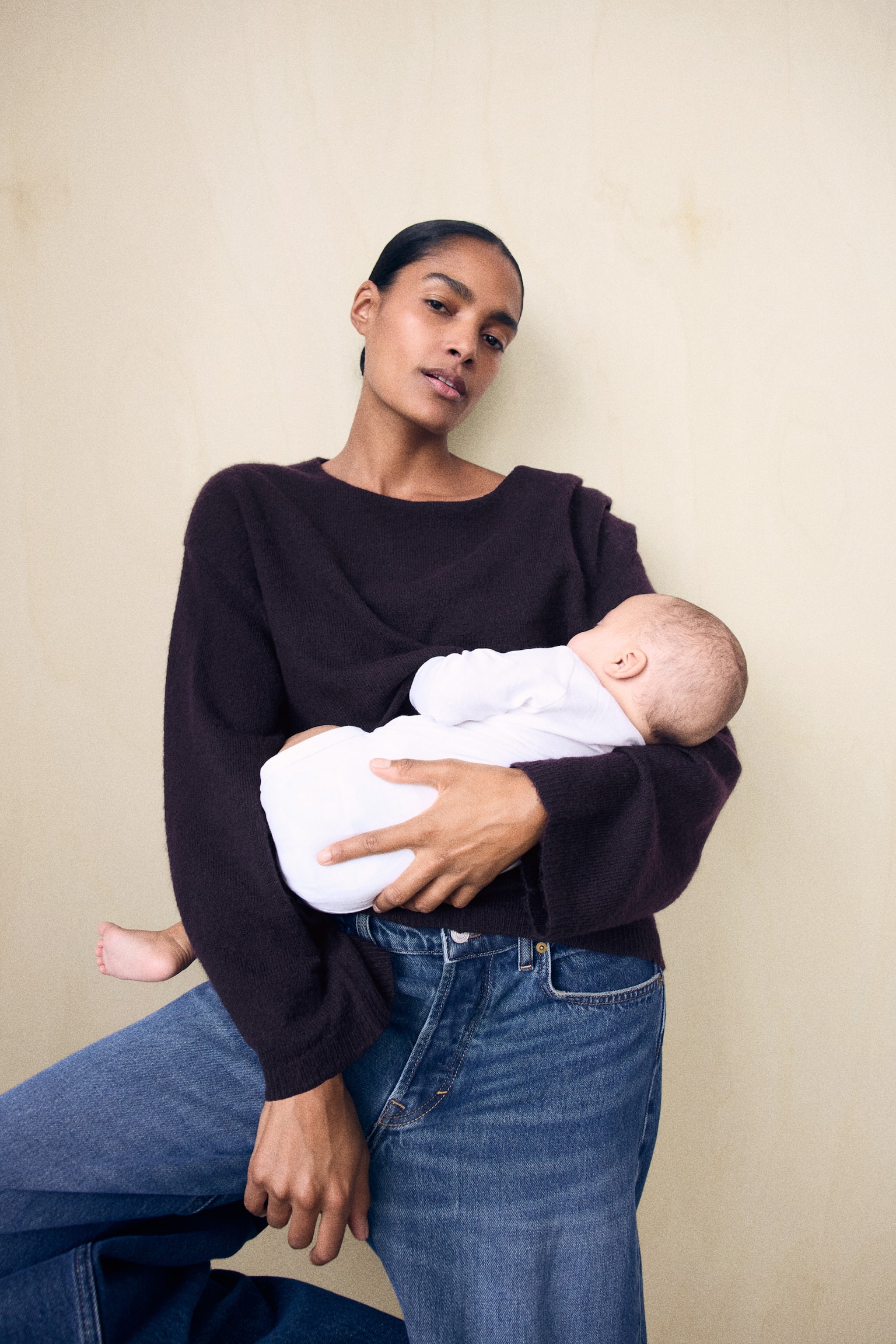 A loose-fit, dark plum knit nursing sweater featuring a round neckline and dropped shoulders is being worn by the model, who is holding a baby dressed in white. The long-sleeved sweater is paired with blue jeans and features ribbed cuffs. - 1