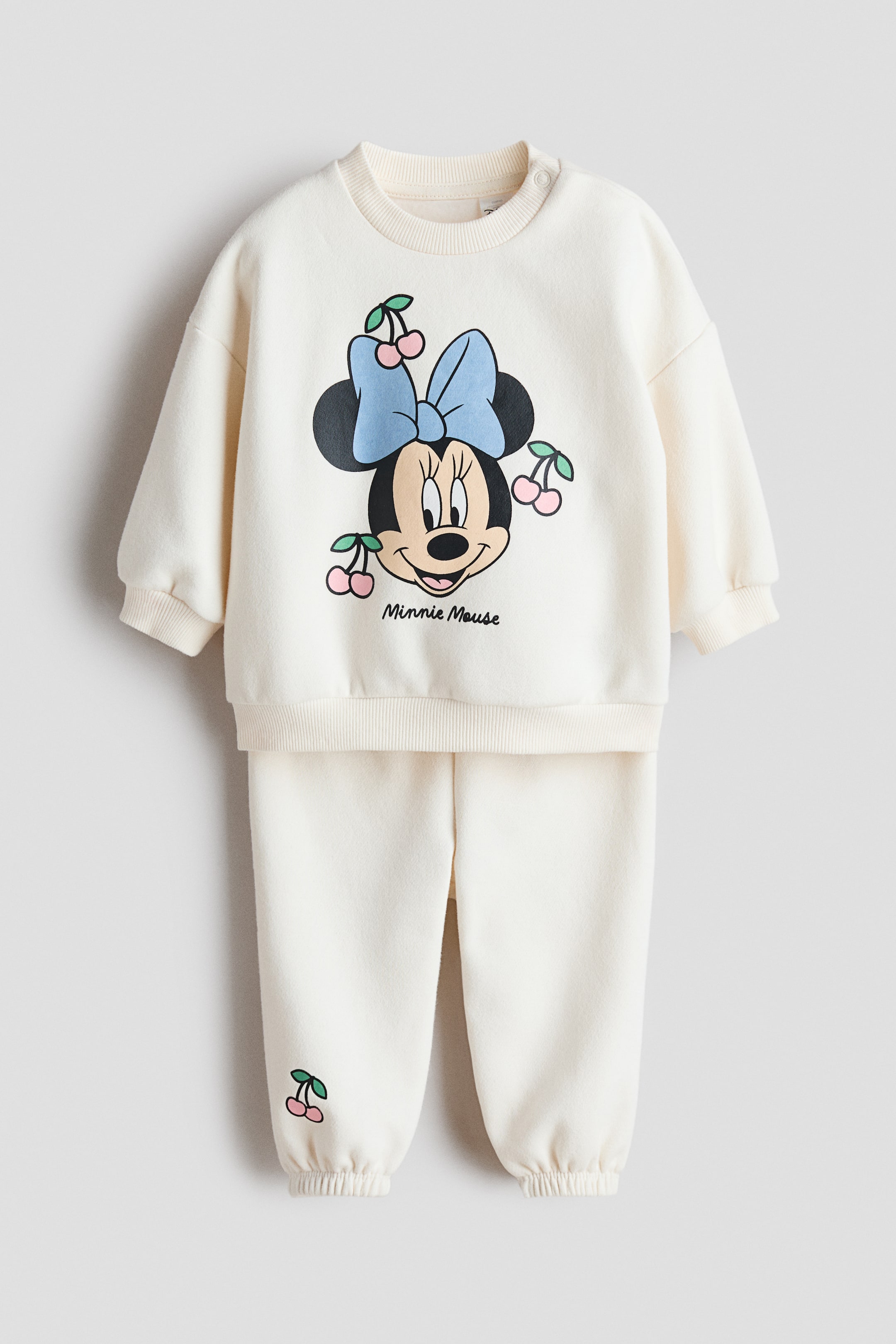 Voir une image plus grande: Un ensemble de survêtement imprimé blanc cassé 2 pièces est présenté face avant. Le sweat-shirt présente un graphique de Minnie Mouse avec un nœud bleu et des cerises roses, un col rond côtelé avec des boutons-pression sur l'épaule, des poignets côtelés et un ourlet côtelé. Le pantalon jogger assorti affiche un petit graphique de cerise sur la jambe gauche et des poignets de cheville côtelés.