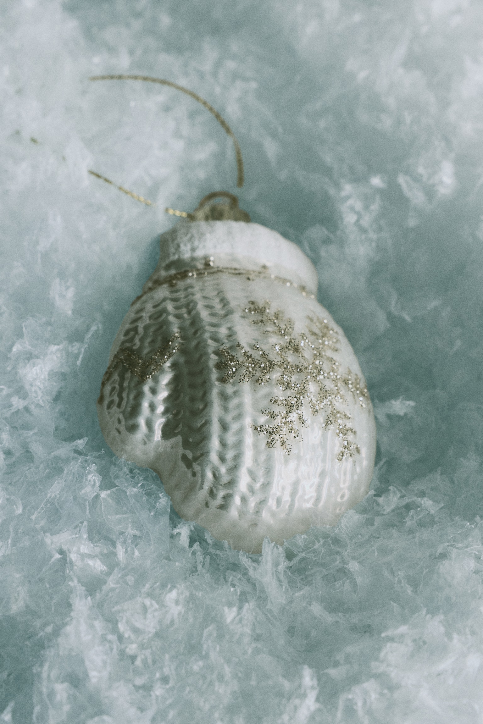 A mitten-shaped glass Christmas ornament faces forward, showcasing a white textured cable-knit pattern and a gold glitter snowflake design on its surface. The ornament features a textured white cuff at the top, complete with a gold-tone hanger loop and a thin gold string. - 1