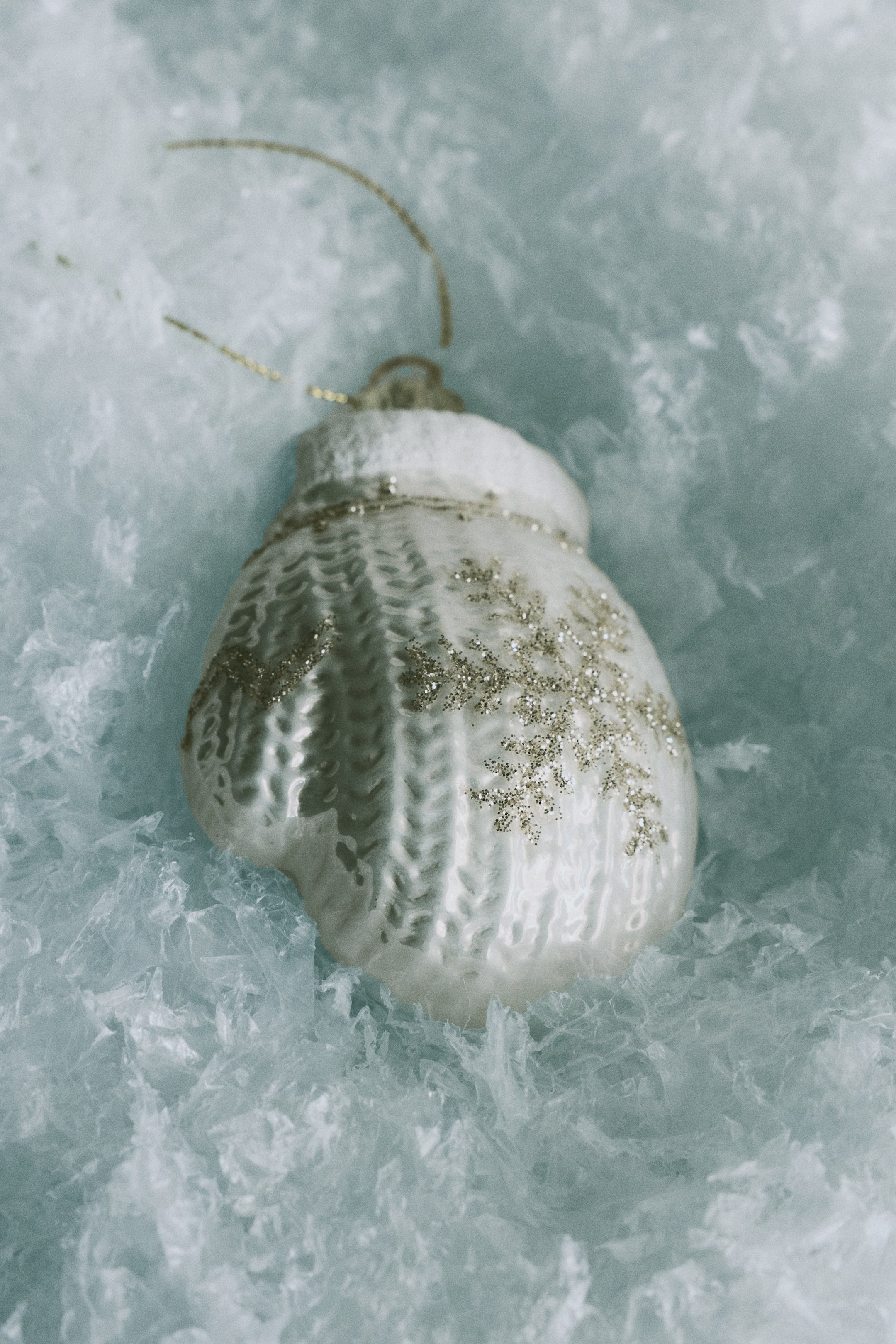 Agrandir l'image: Un ornement de Noël en verre en forme de moufle est présenté de face, mettant en valeur un motif torsadé texturé blanc façon tricot et un design flocon de neige scintillant doré sur sa surface. L’ornement présente un revers texturé blanc en partie supérieure, complété par une boucle de suspension ton or et un fin cordon doré.
