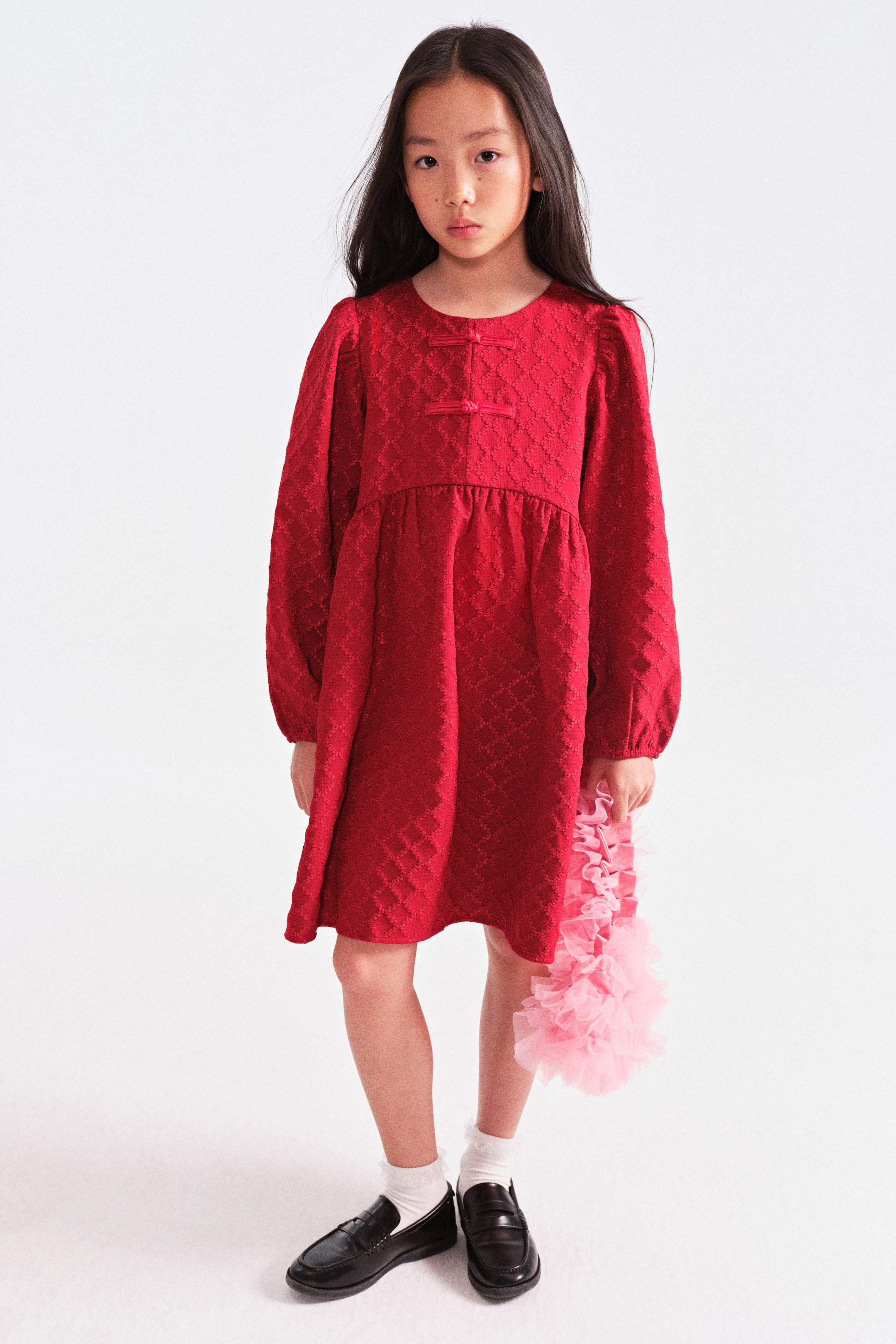 View larger image: Facing forward, the red long-sleeved dress showcases a raised diamond pattern across the fabric. It features a round neckline with two decorative red frog closures on the upper front of the bodice, and falls into a gathered, flared skirt from an empire waistline.