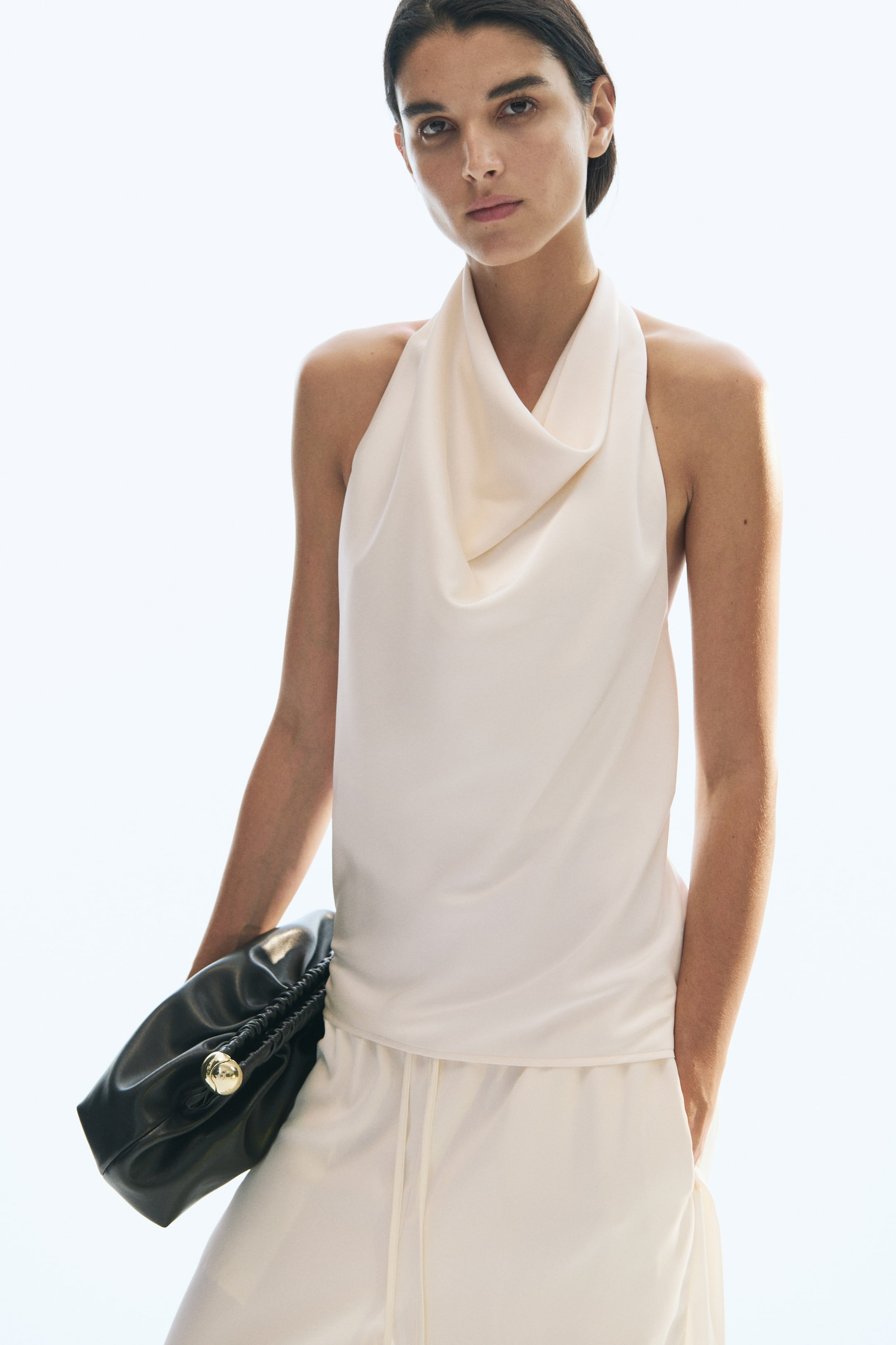 View larger image: A sleeveless, loose-fit halterneck top in cream draping satin is worn by the model, featuring a draped cowl neckline. The top is styled with matching cream trousers, while a black leather clutch bag is tucked under the model's arm.
