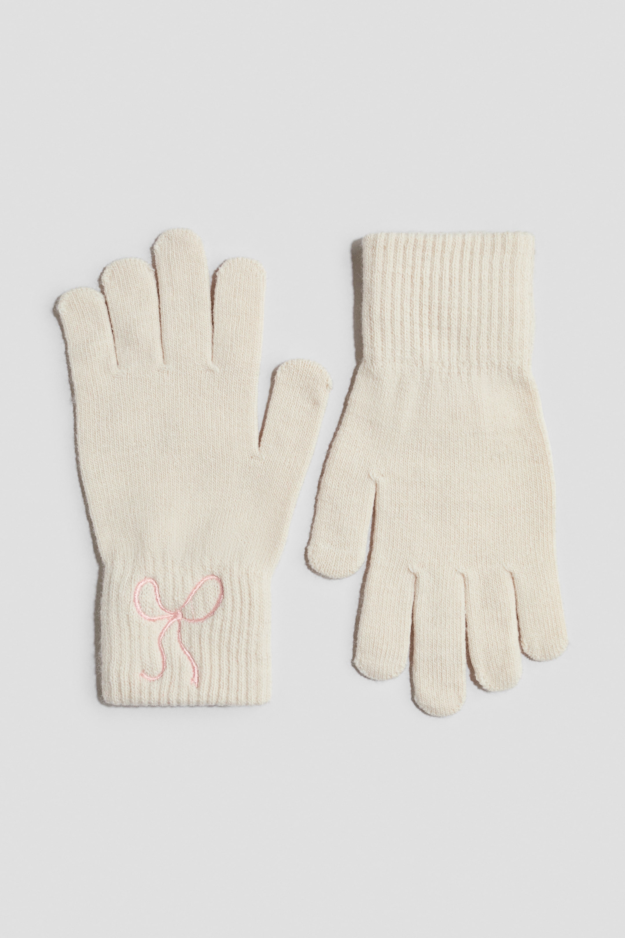 Grösseres Bild ansehen: Ein Paar cremefarbene Strickhandschuhe liegt flach da, wobei ein Handschuh mit der Vorderseite nach oben liegt, um eine kleine, bestickte rosa Schleife an seinem gerippten Bündchen zu zeigen. Der zweite Handschuh ist mit den Fingern nach unten ausgerichtet und zeigt das saubere, fein gestrickte Design sowie das gerippte Bündchen.