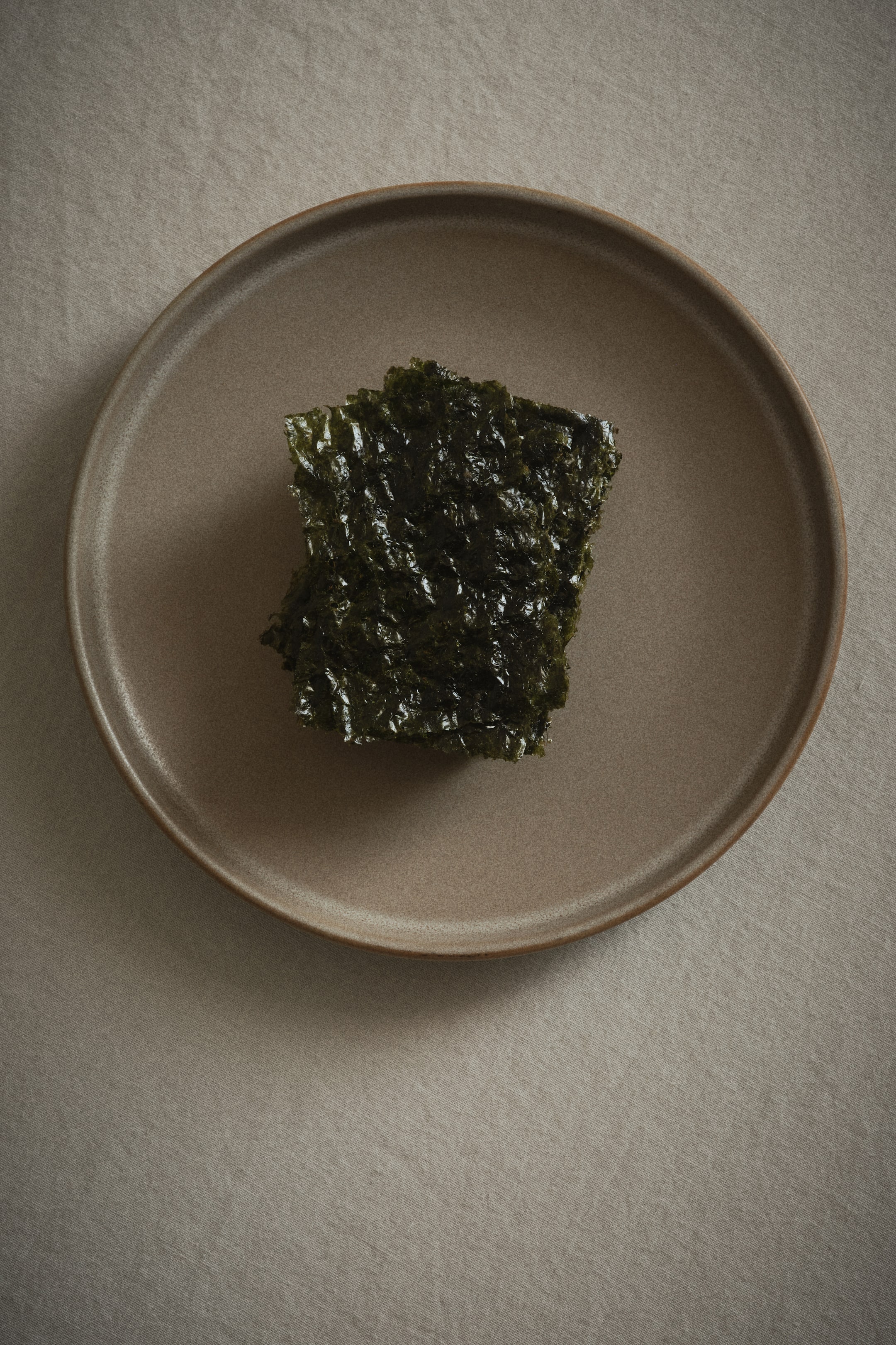 View larger image: A stack of dark green, crinkled roasted seaweed sheets is resting centrally on a round, flat, beige-brown plate, presented from a top-down perspective.
