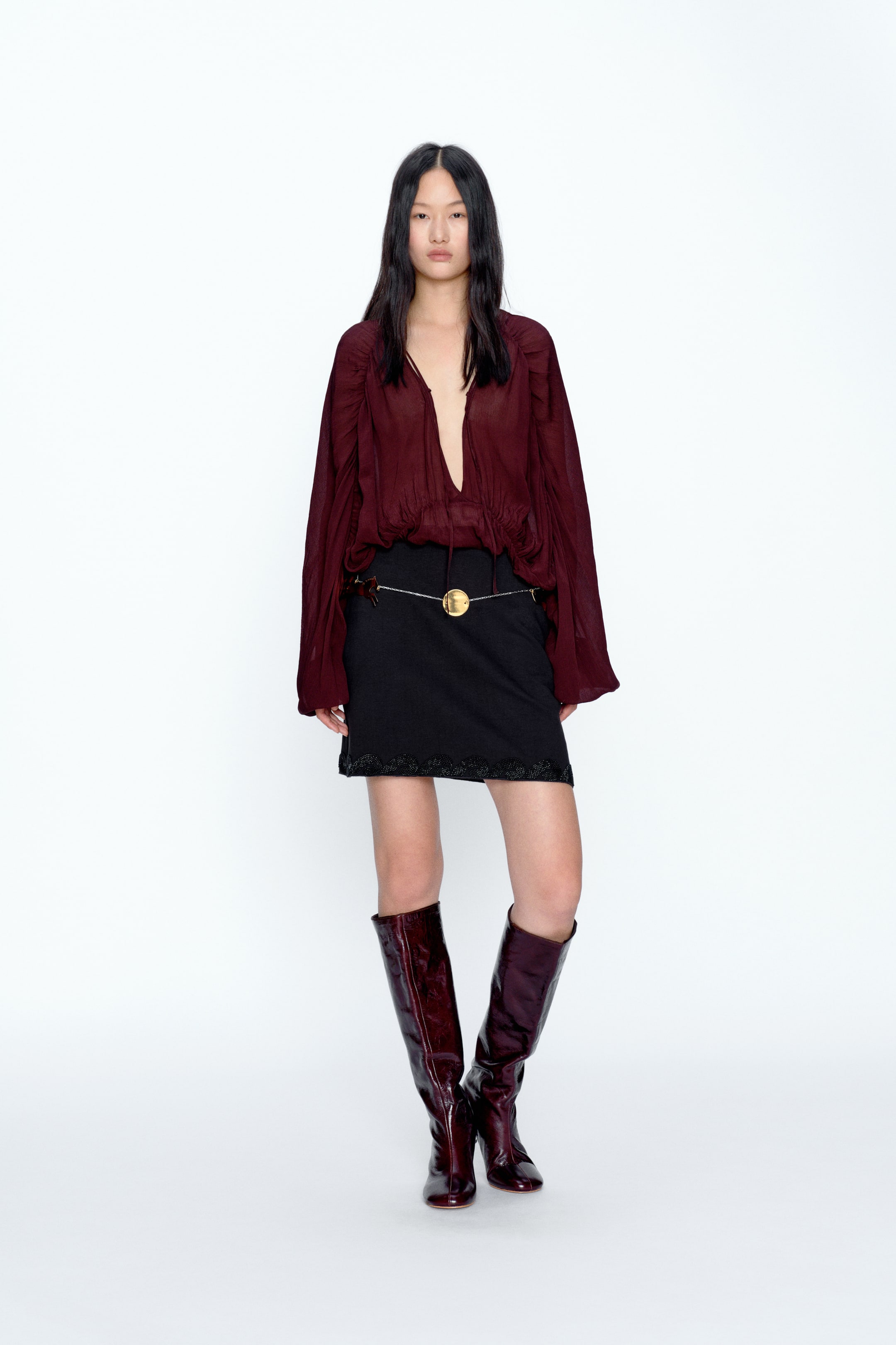 View larger image: A maroon blouse featuring a deep V-neckline with a tie closure and voluminous long sleeves gathered at the shoulders and cuffs. It is paired with a black mini skirt with a decorative hem, cinched with a gold chain belt with a central medallion, and styled with dark maroon glossy knee-high boots.