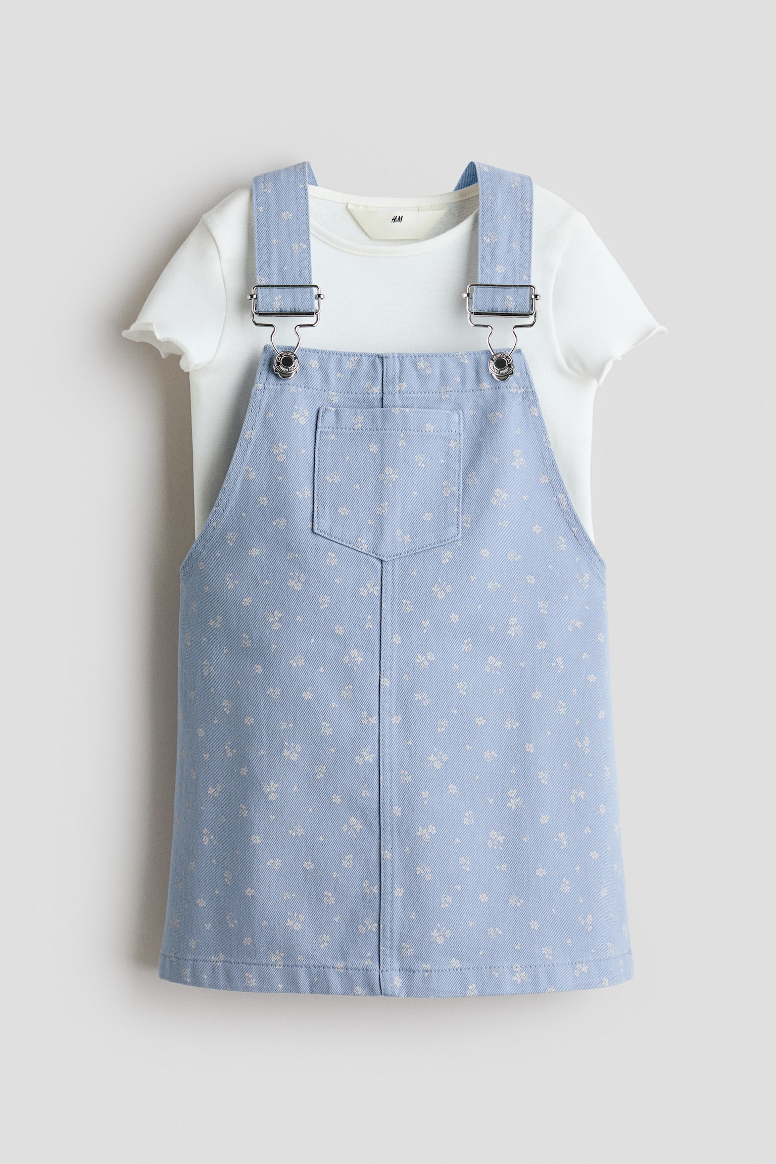 This set is featuring a white short-sleeved top in soft cotton jersey with ruffled edges at the sleeves, layered under a light blue dungaree dress in lightweight cotton twill, facing front. The dungaree dress is displaying a small white floral print, a chest pocket, and adjustable straps with metal fasteners.