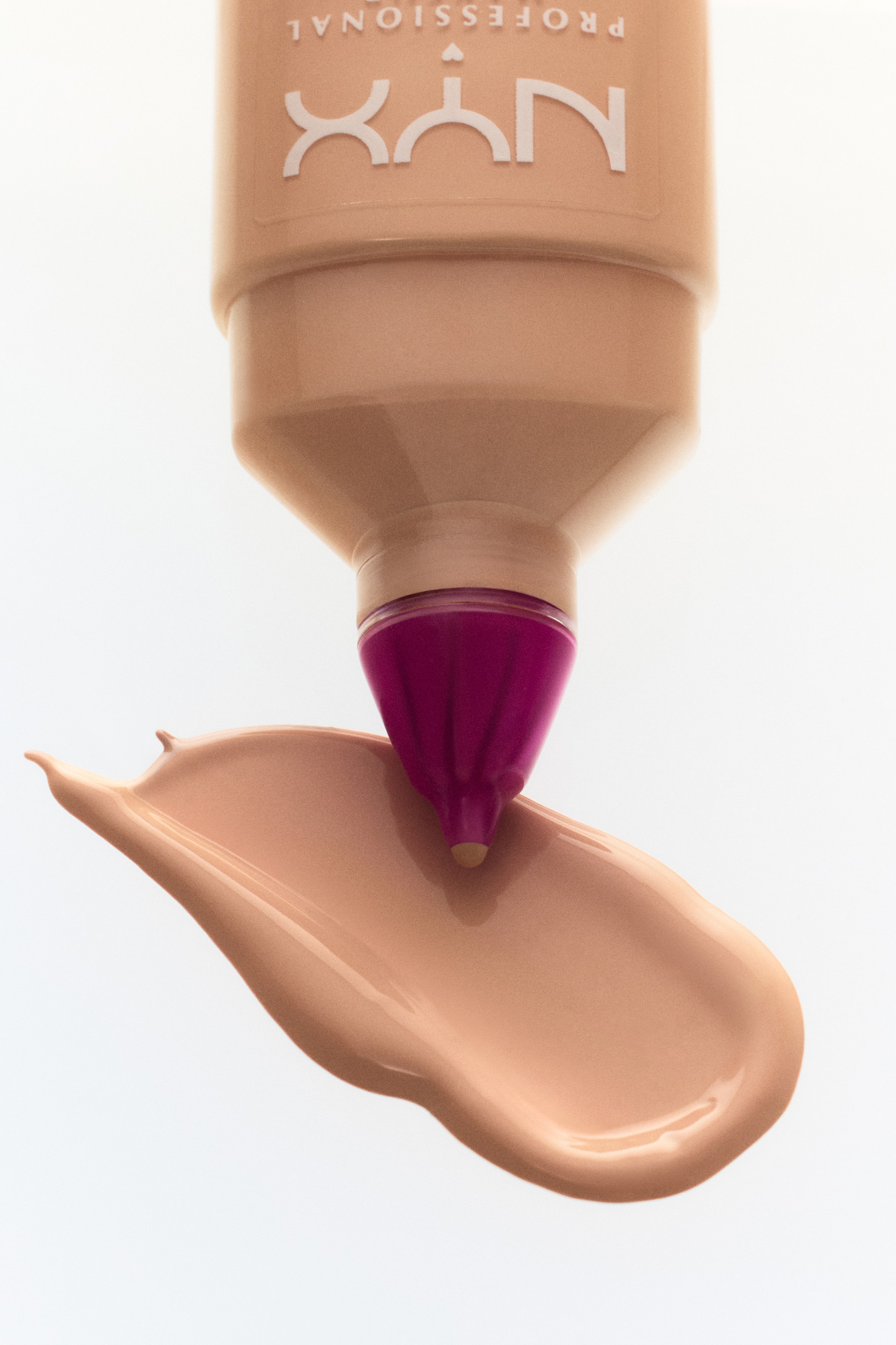 Vis større bilde: En lys beige NYX PROFESSIONAL foundationflaske, med hvit skrift som vises opp ned, dispenserer en jevn, lys beige flytende foundation. Den magenta, ribbede applikatorspissen berører den tykke strimmelen av foundation.