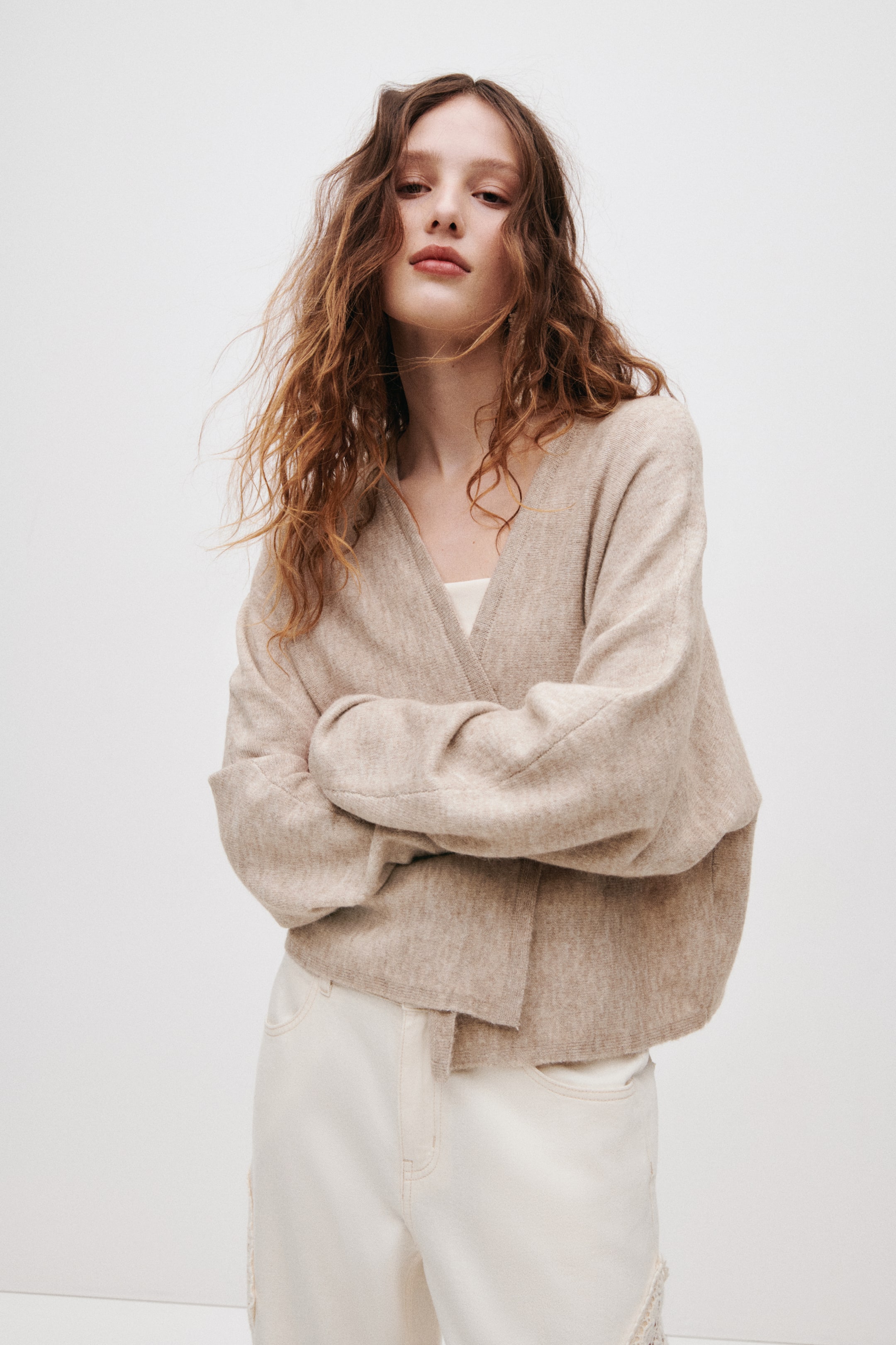 Visa större bild: En beige stickad omlottkofta presenteras, med en djup V-ringning och nedhasade axlar. Dess avslappnade passform visar upp långa ärmar och en omlottfront, med en vit topp synligt lager på lager under.