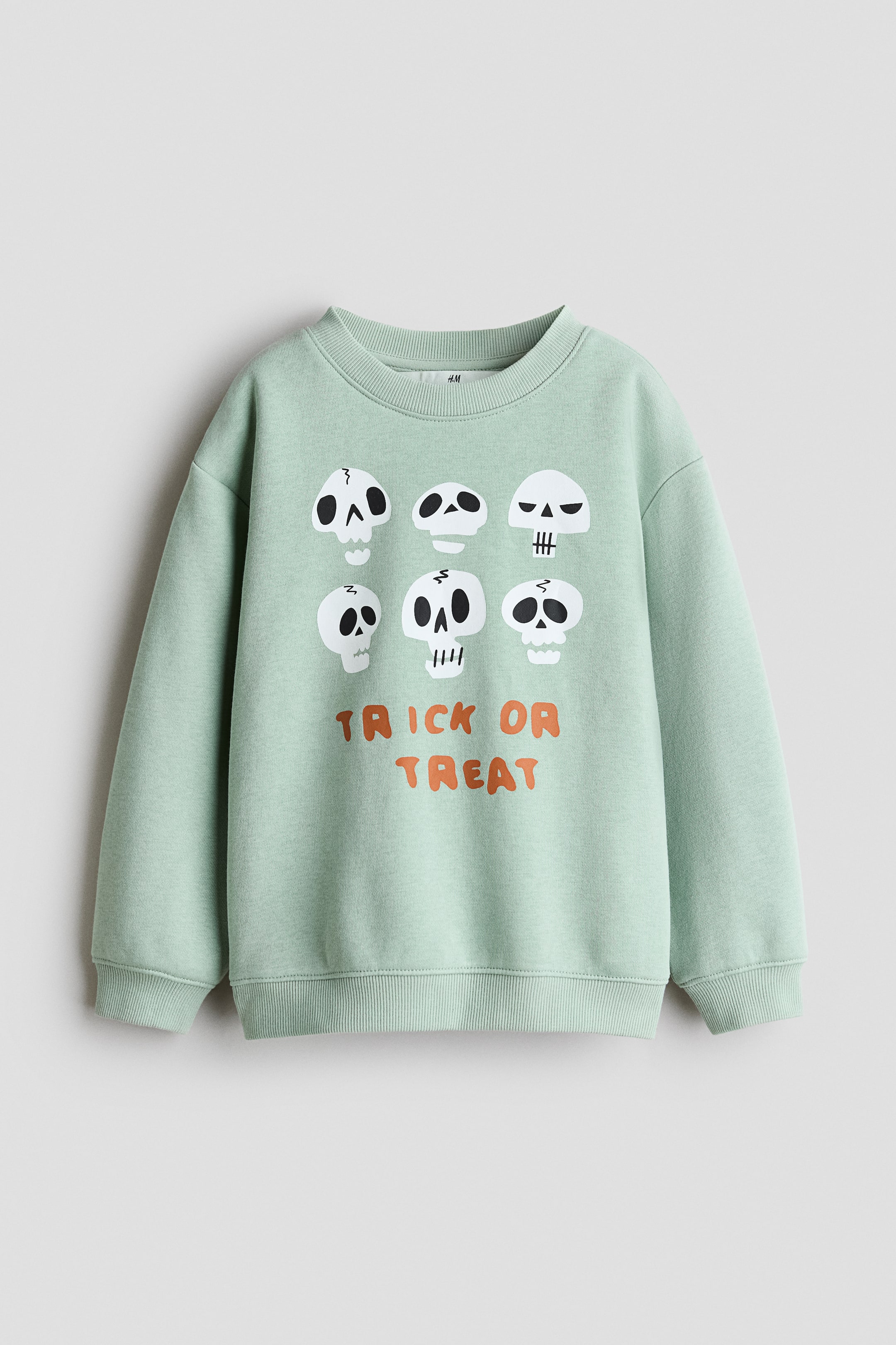 View larger image: A light green sweatshirt is showcasing its front, featuring a round, rib-trimmed neckline, dropped shoulders, and long sleeves with ribbed cuffs. The hem is also ribbed, and six white skull graphics are decorating the upper front above the orange text 'TRICK OR TREAT'.